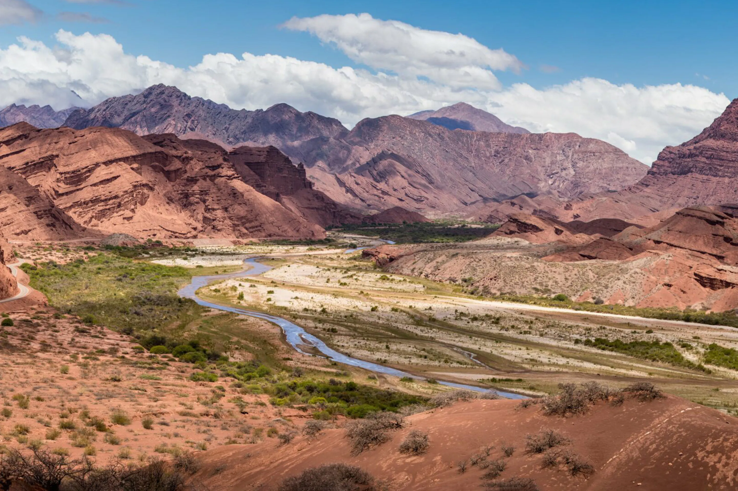 A winding river runs through red rock valleys and layered mountains in northwest Argentina beneath bright clouds.
