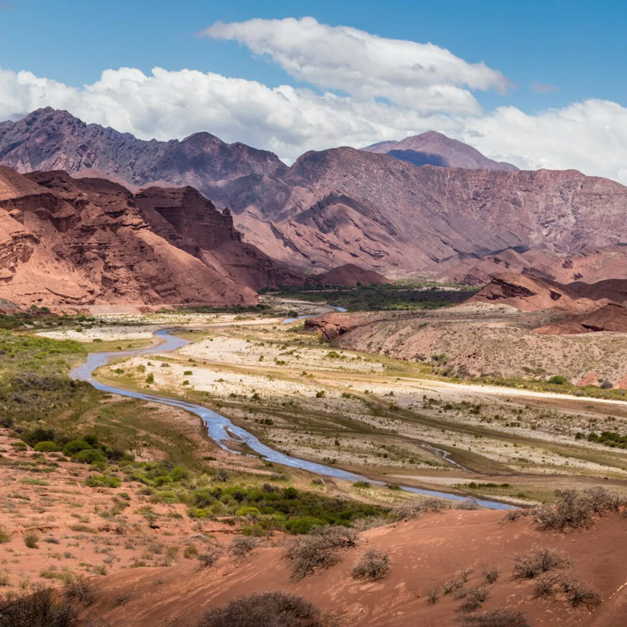 A winding river runs through red rock valleys and layered mountains in northwest Argentina beneath bright clouds.