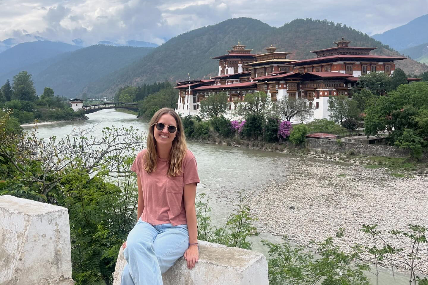 A traveler sits before Punakha Dzong, with red roofs, white walls,  and Bhutan's river valley around her.