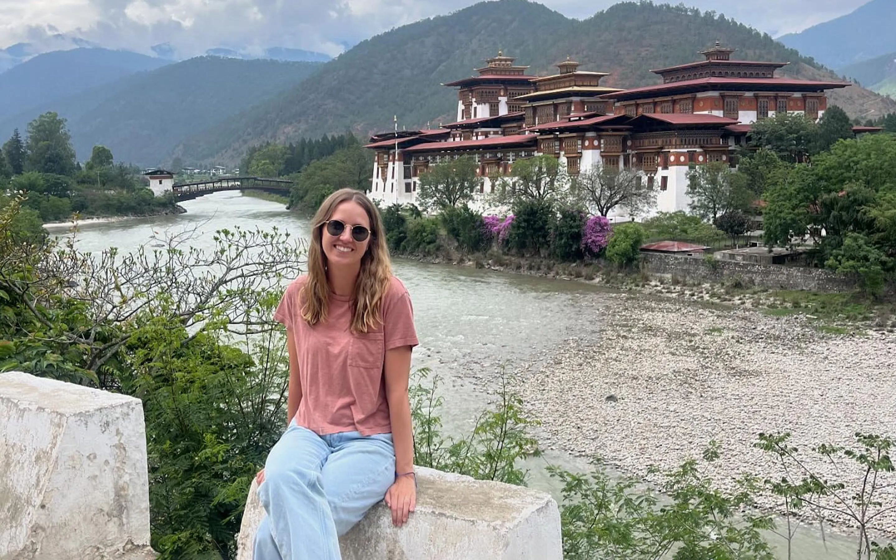 A traveler sits before Punakha Dzong, with red roofs, white walls, and Bhutan's river valley around her.