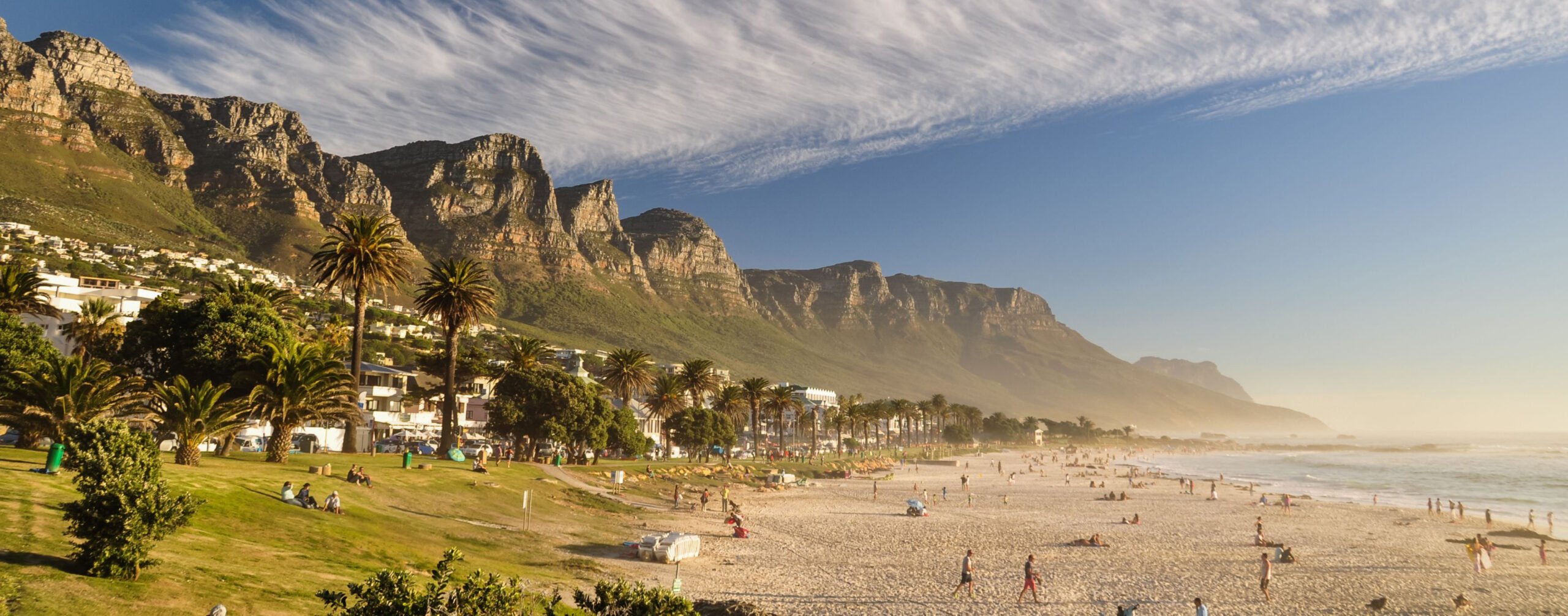 Camps Bay beach stretches beneath the Twelve Apostles near Cape Town, with pale sand and waves in the foreground.