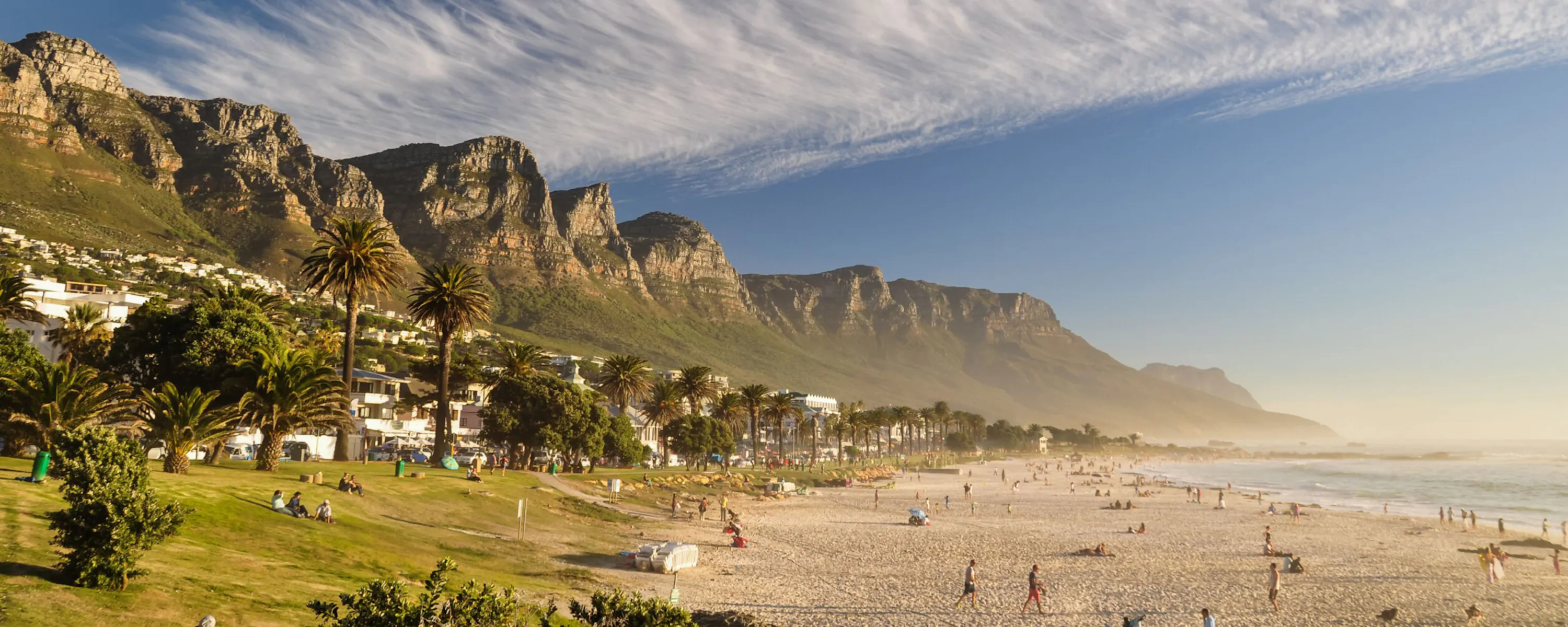 Camps Bay beach stretches beneath the Twelve Apostles near Cape Town, with pale sand and waves in the foreground.