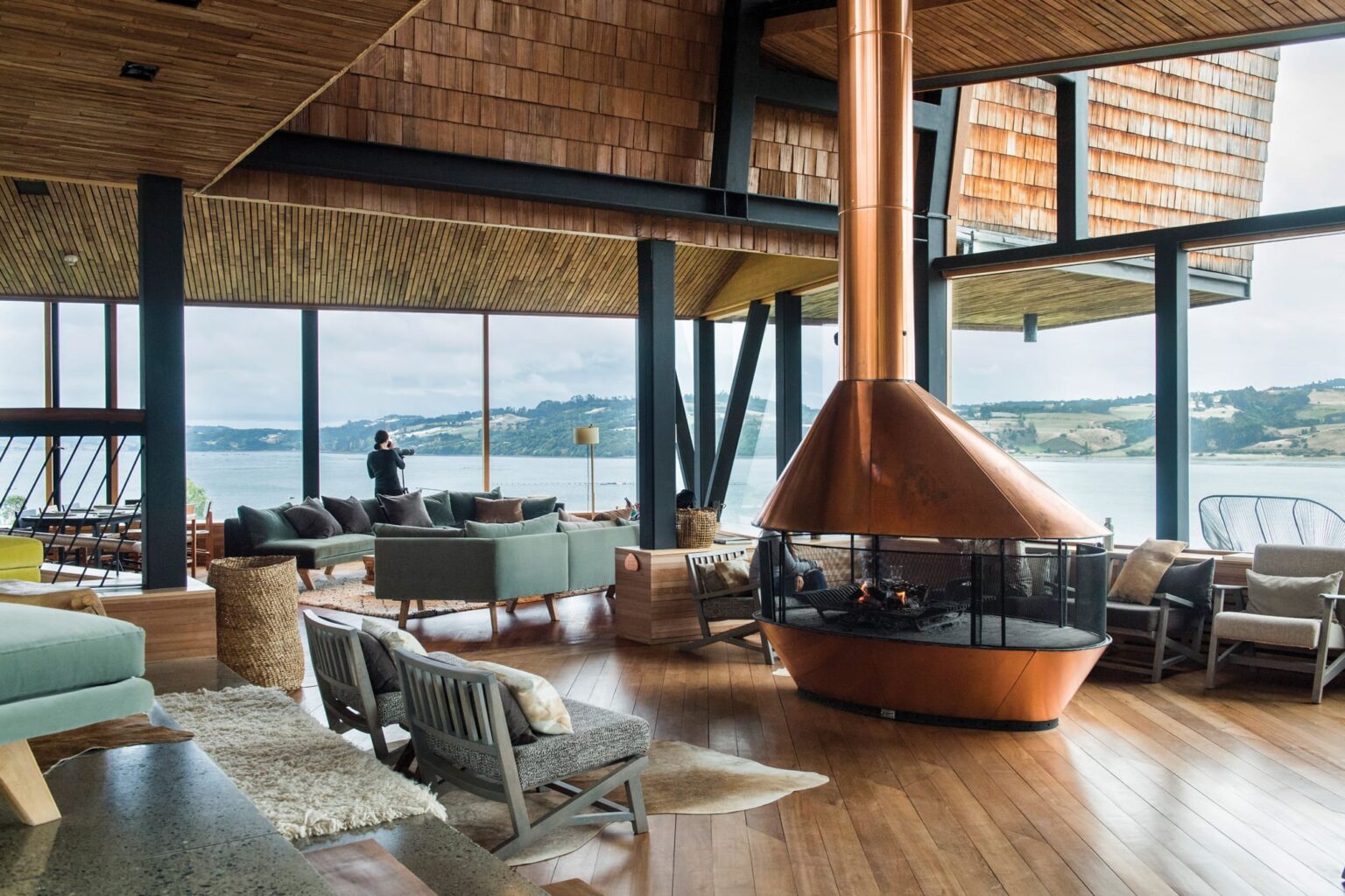 A timber lodge lounge with fireplace and wide water views at Tierra Chiloe, set in Chile's Chiloe Island.