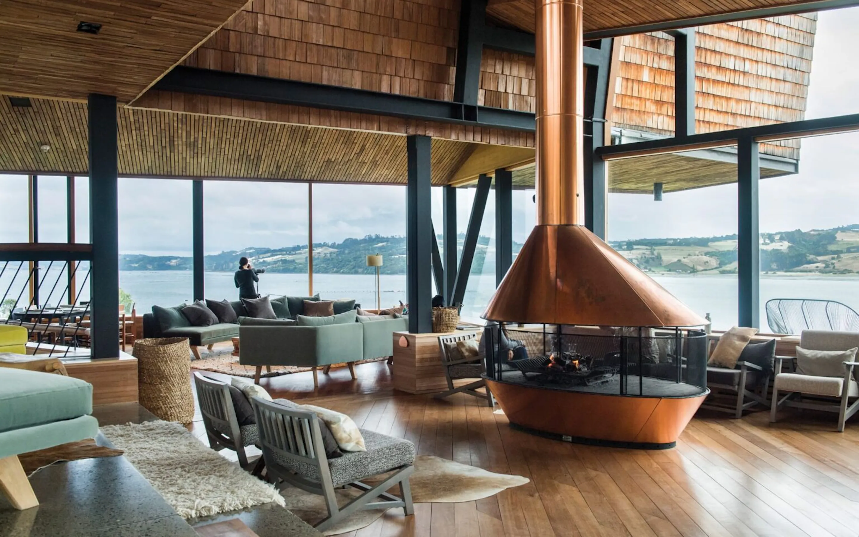 A timber lodge lounge with fireplace and wide water views at Tierra Chiloe, set in Chile's Chiloe Island.