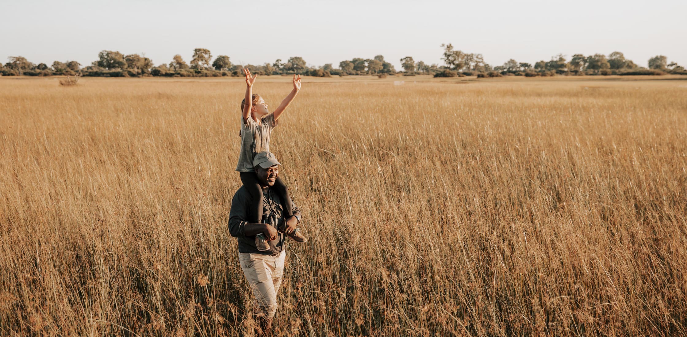 A child stands in tall Okavango grass, holding a branch high while a guide watches nearby under open sky.