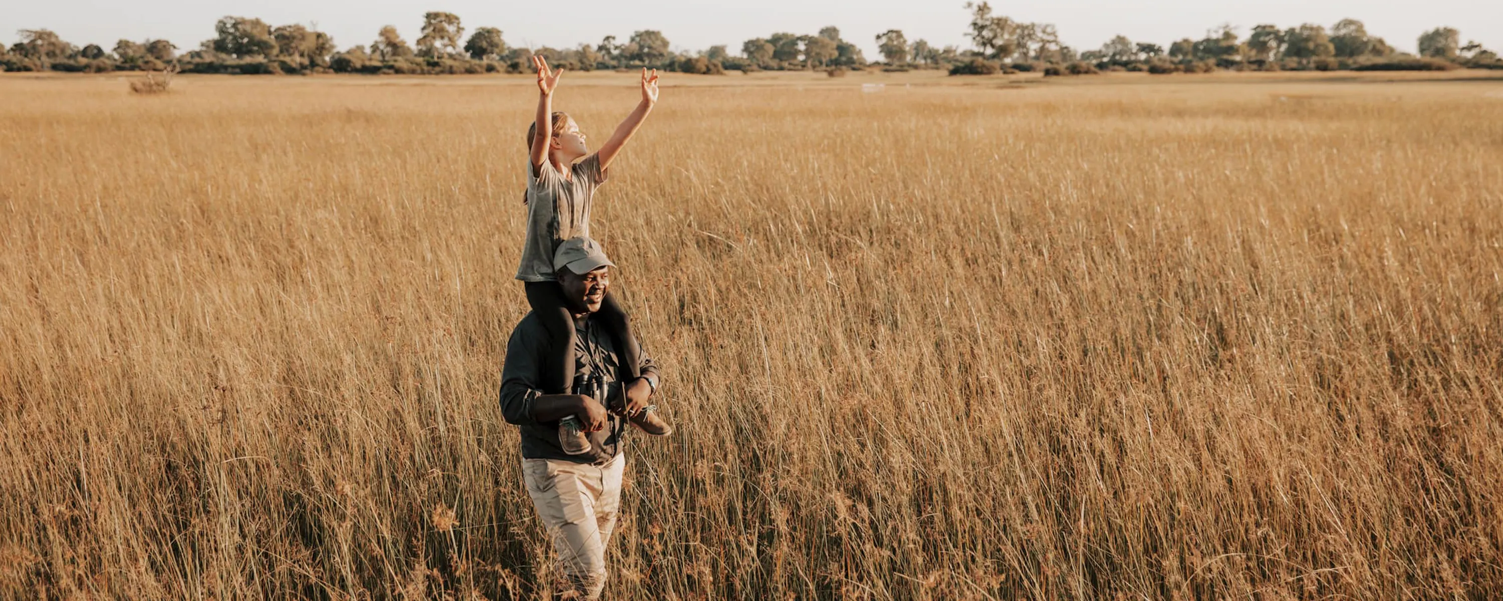 A child stands in tall Okavango grass, holding a branch high while a guide watches nearby under open sky.