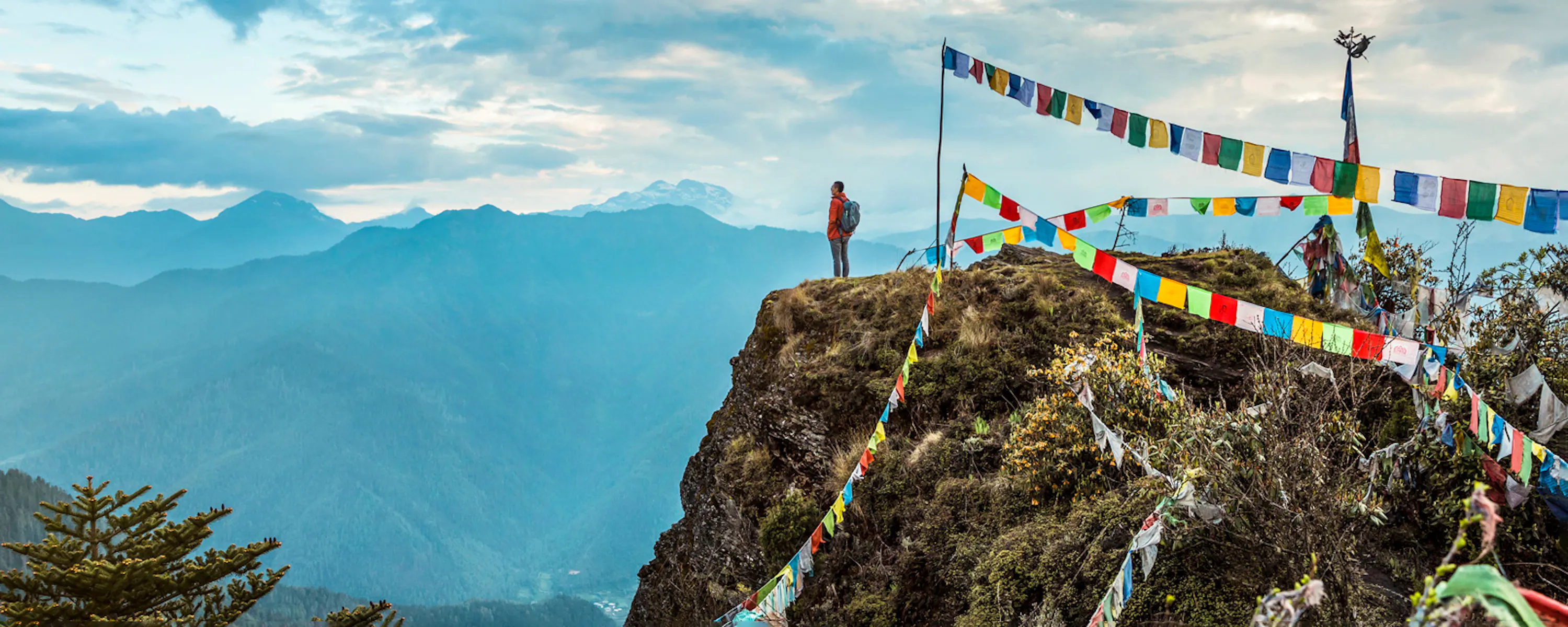 Prayer flags flutter from a rocky peak above COMO Uma Paro, with layers of blue mountains filling the distance.