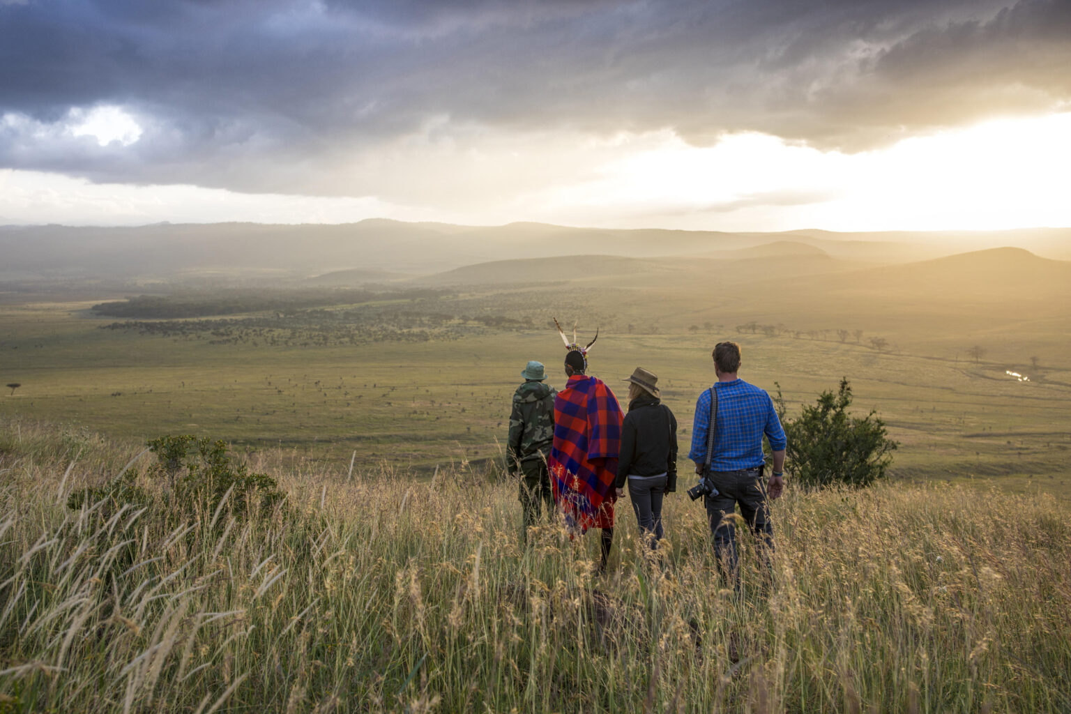 Hikers cross open green fields near Sirikoi Lodge, with forested hills rising beyond Lewa in Kenya.