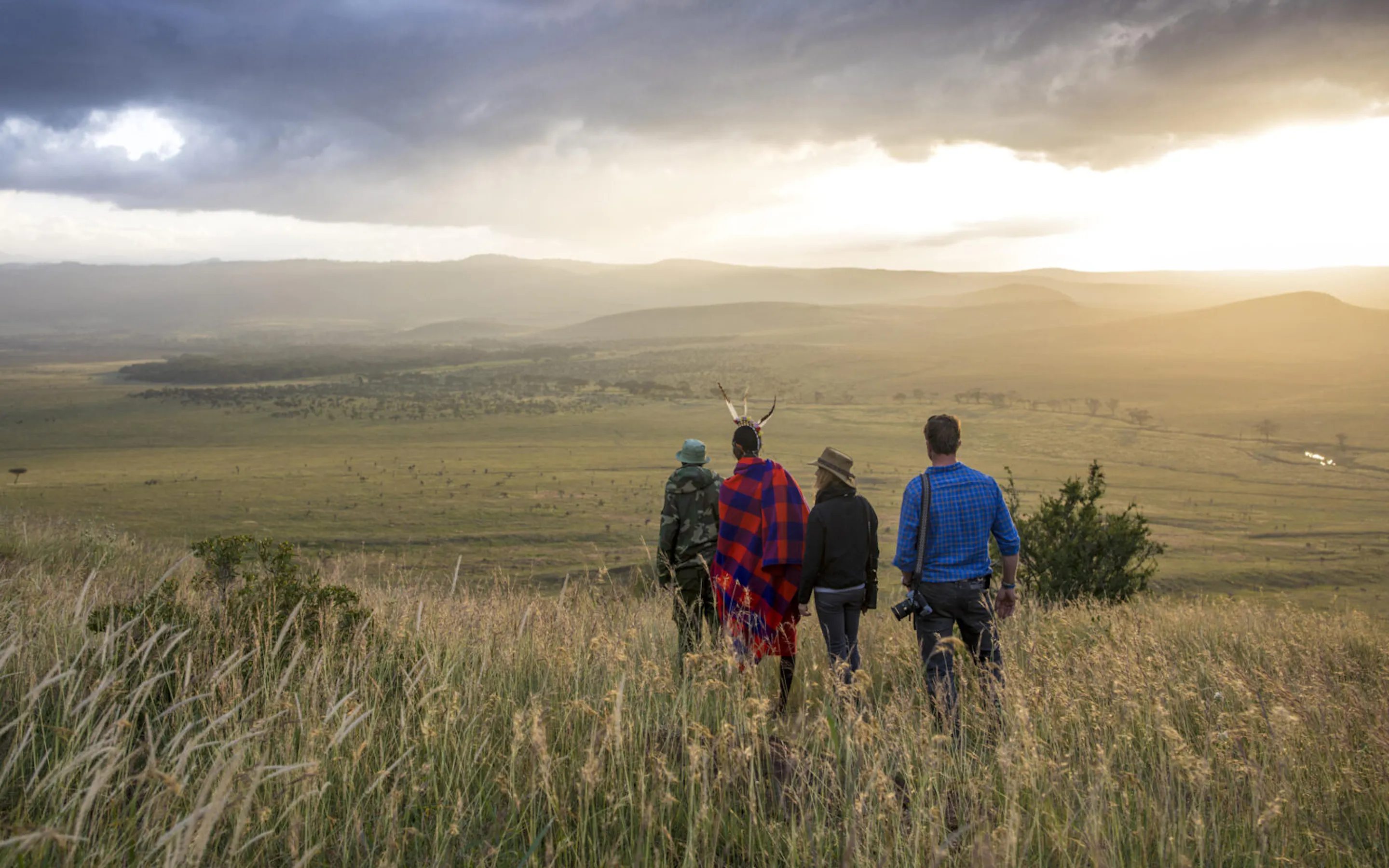 Hikers cross open green fields near Sirikoi Lodge, with forested hills rising beyond Lewa in Kenya.