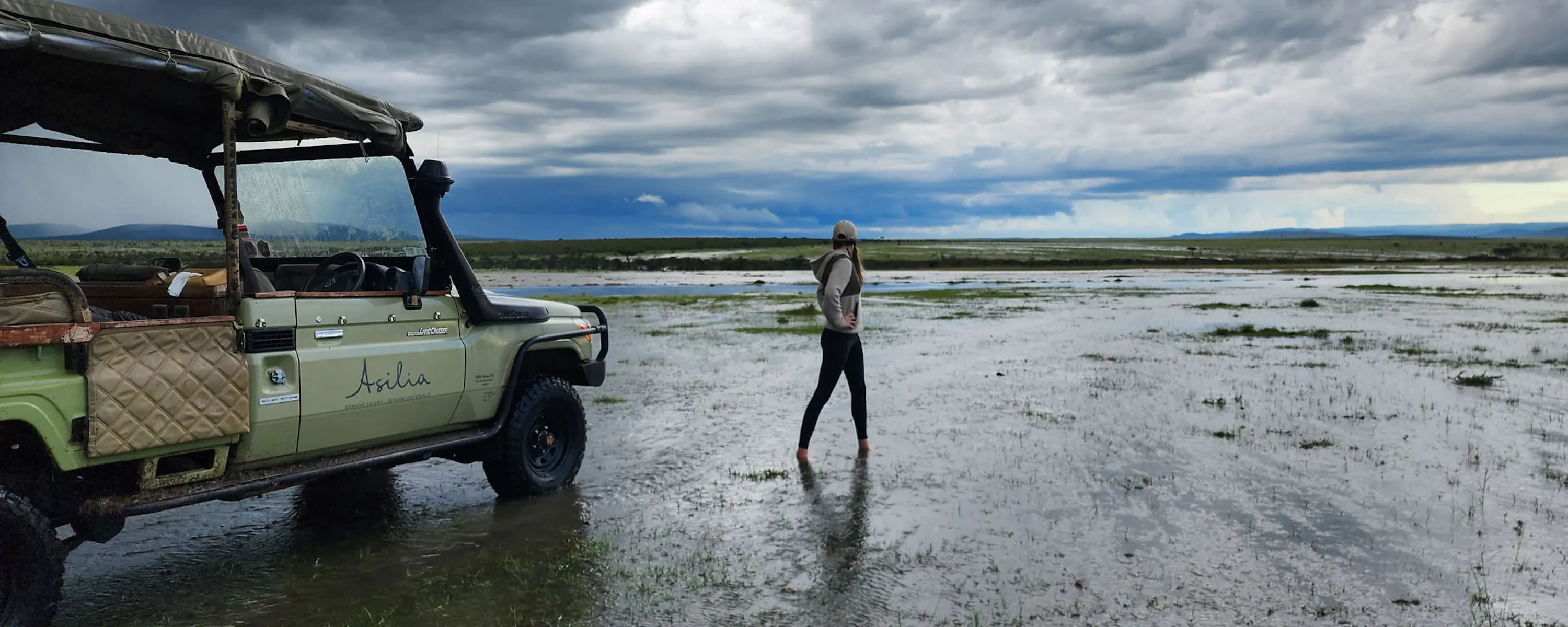 A traveler standing beside a safari vehicle on flooded plains against the backdrop of Kenya's Maasai Mara.