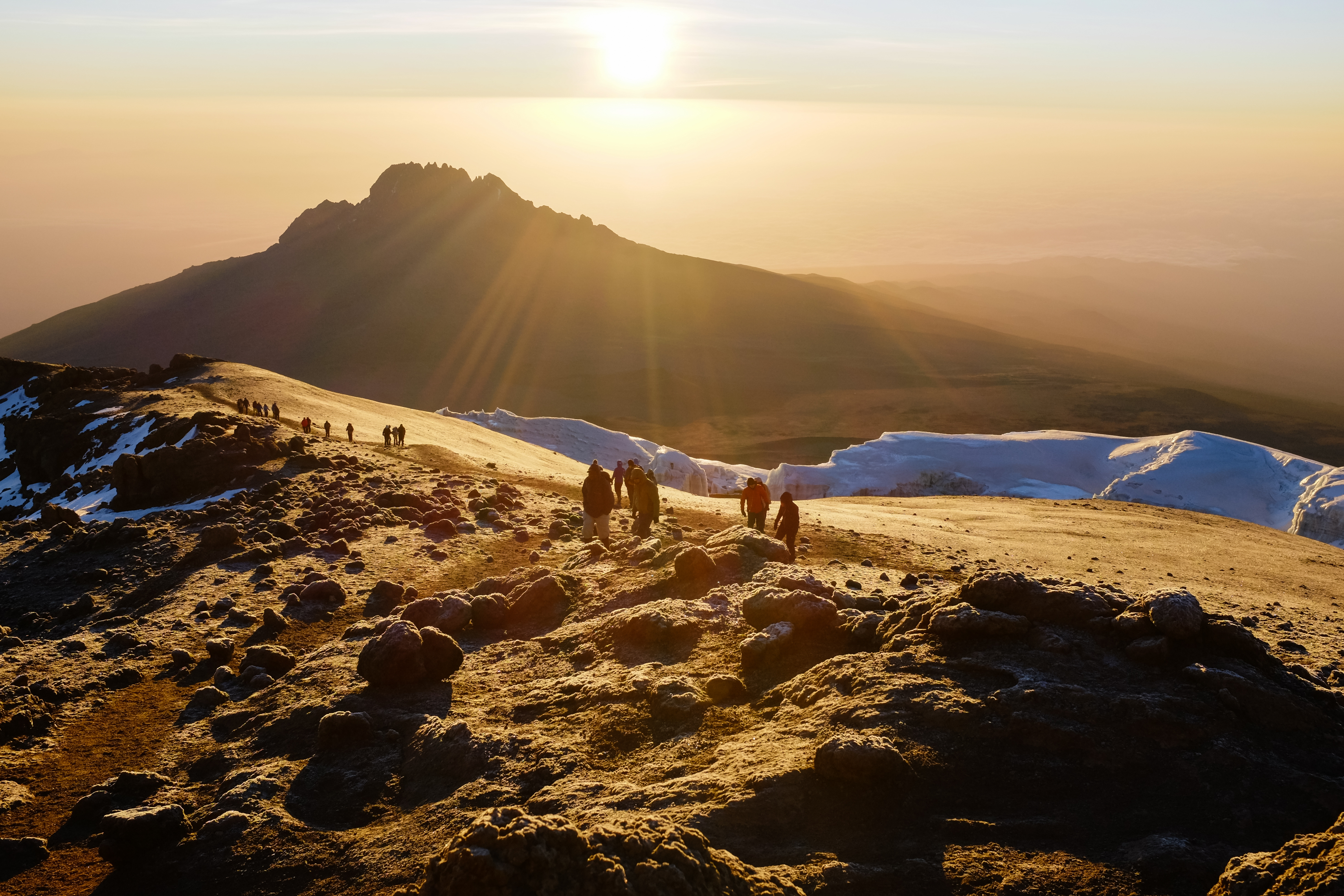 Trekkers stand on a ridge at sunrise on Kilimanjaro, with warm light spilling over snow and a distant summit.