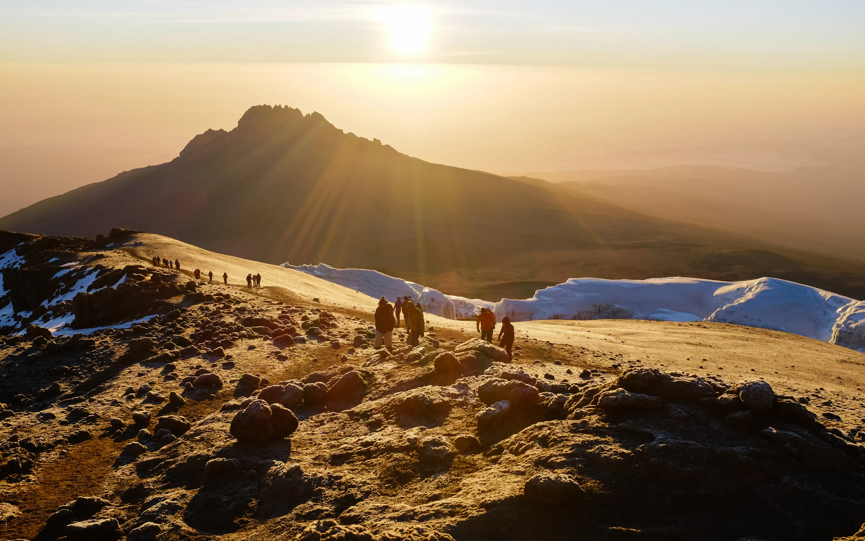 Trekkers stand on a ridge at sunrise on Kilimanjaro, with warm light spilling over snow and a distant summit.