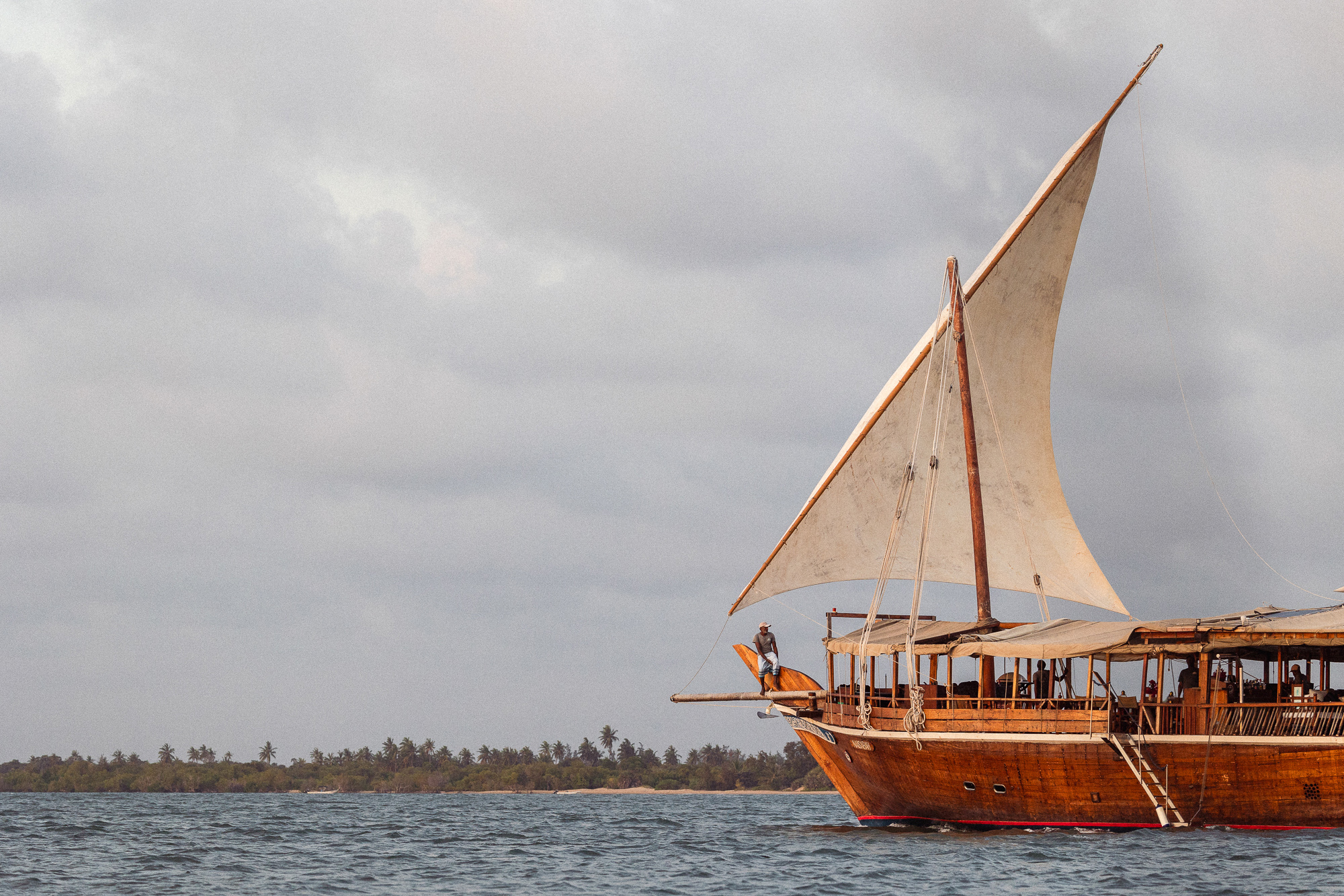 A traditional dhow sails off Lamu beneath moody clouds, its wooden hull and tall mast rising above the sea.