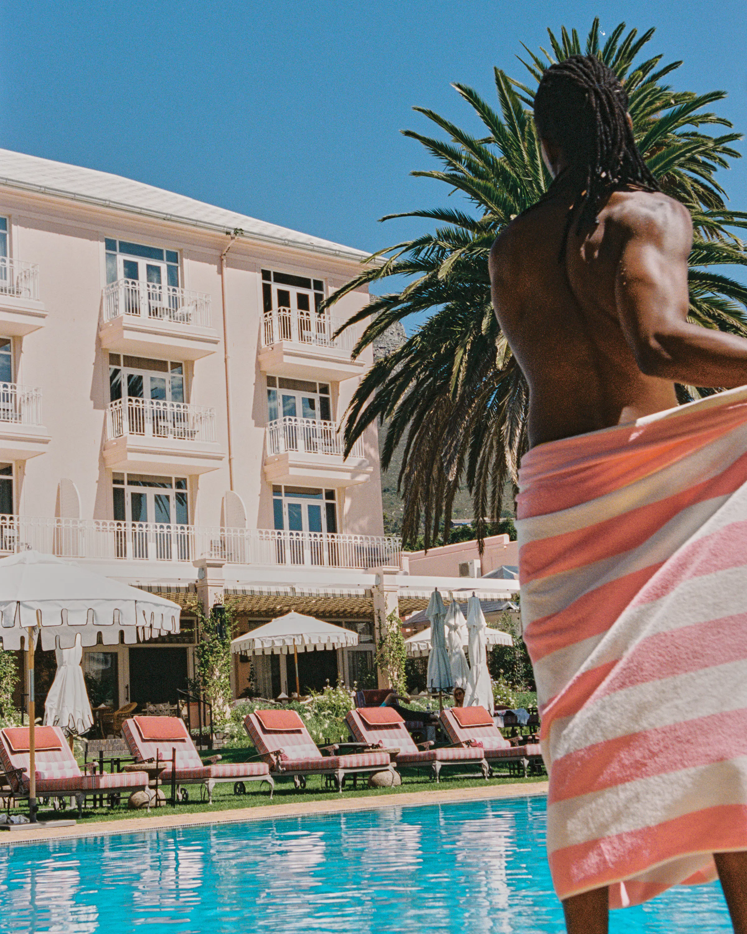 A guest wrapped in a striped towel stands by the Mount Nelson pool, with palms, loungers, and white facades behind.