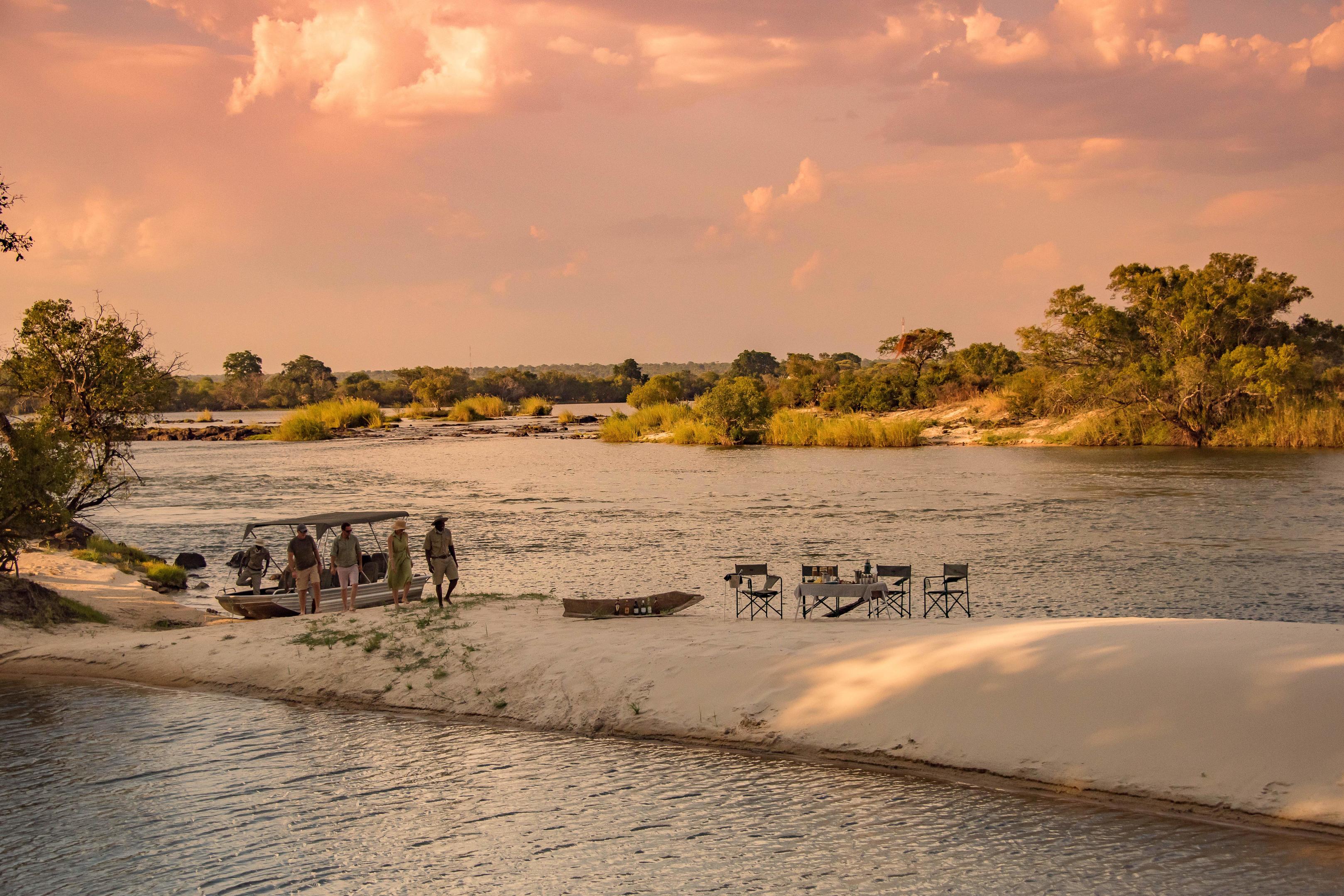 A riverside dinner is set on a sandy bank at Mpala Jena, with calm water and glowing sunset clouds beyond.