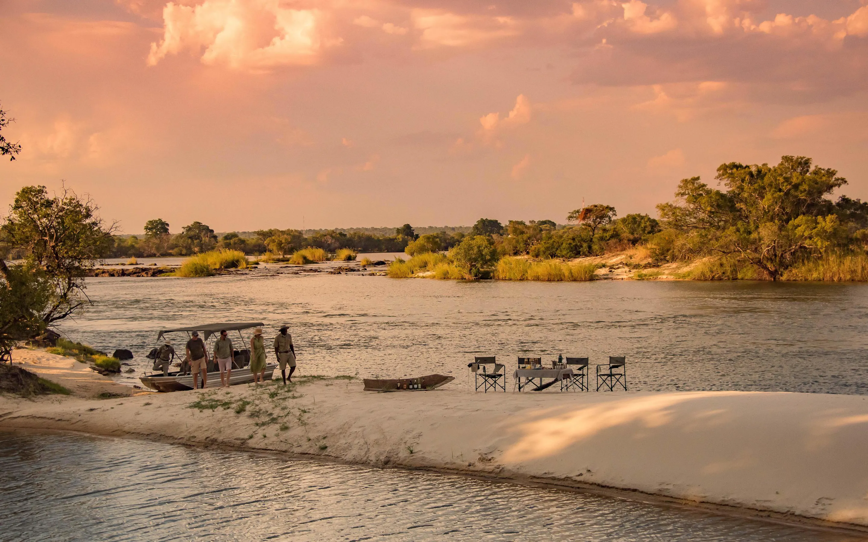 A riverside dinner is set on a sandy bank at Mpala Jena, with calm water and glowing sunset clouds beyond.