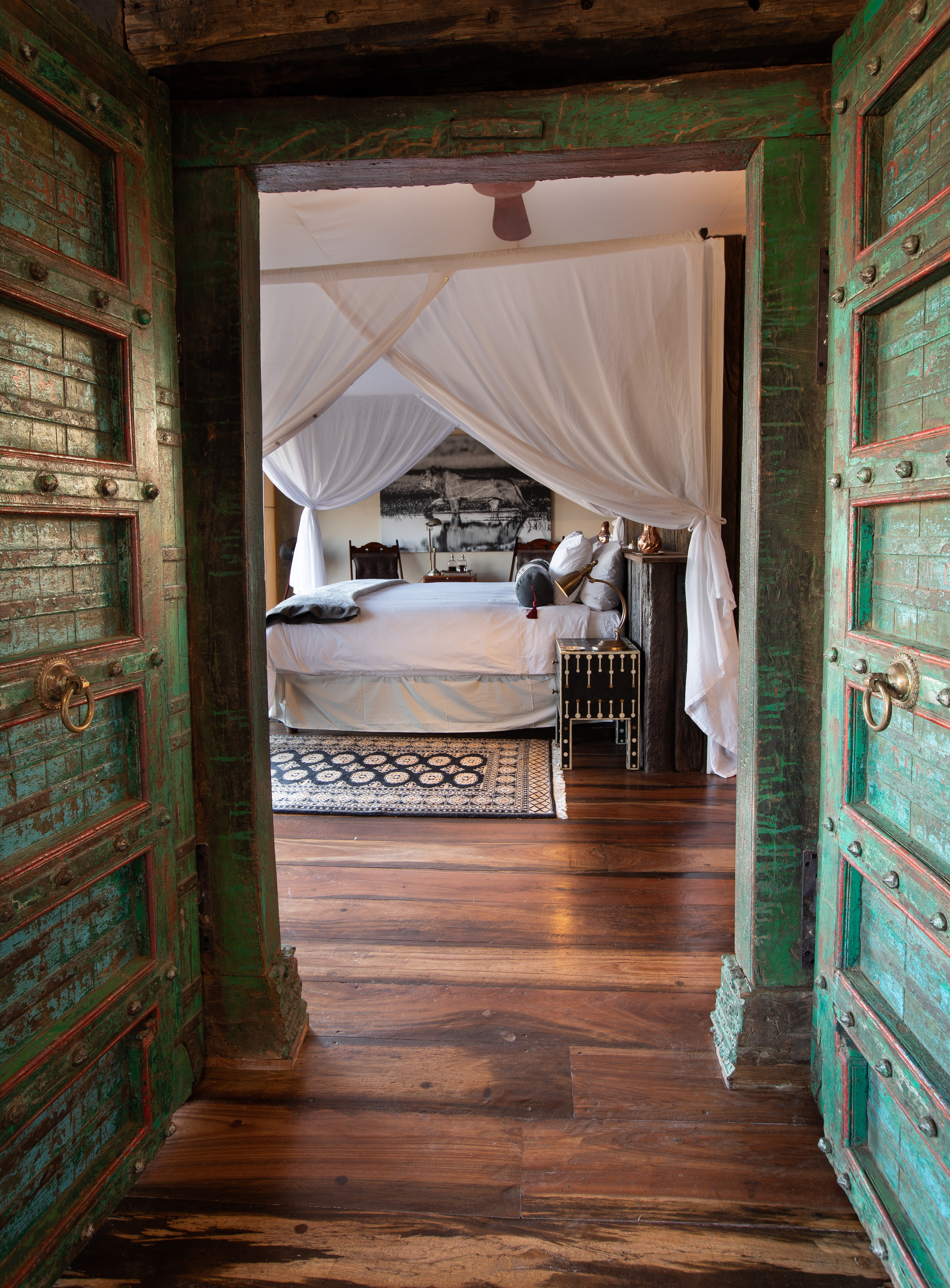 A canopied bedroom at Selinda Camp is seen through weathered doors, with polished floors and soft morning light.