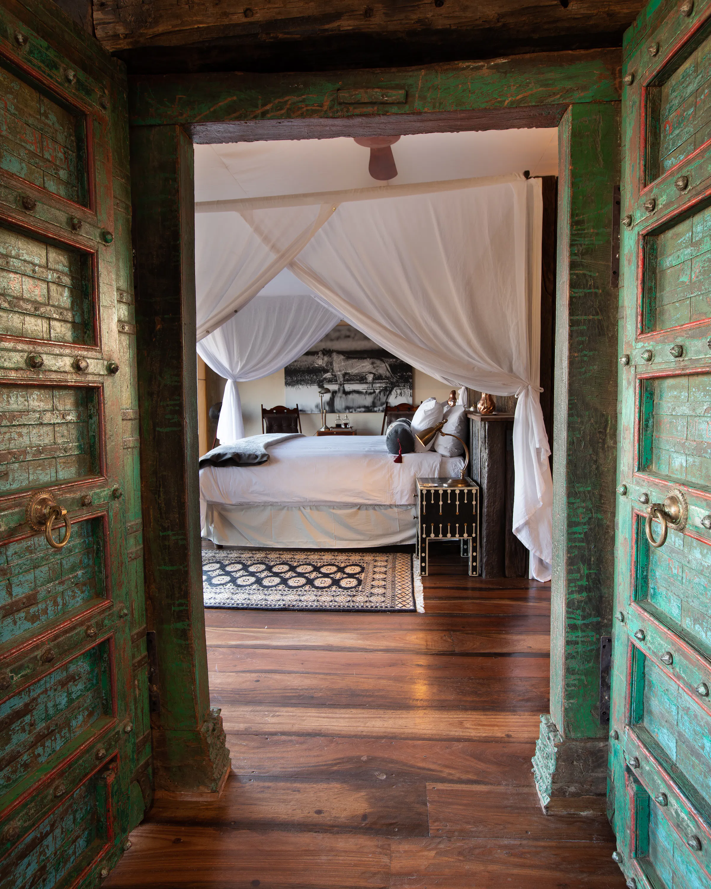 A canopied bedroom at Selinda Camp is seen through weathered doors, with polished floors and soft morning light.