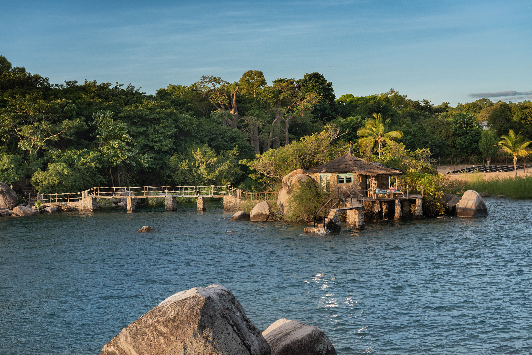 A lakeside pavilion sits above the water at Kaya Mawa, with boulders, palms, and lush island greenery behind.