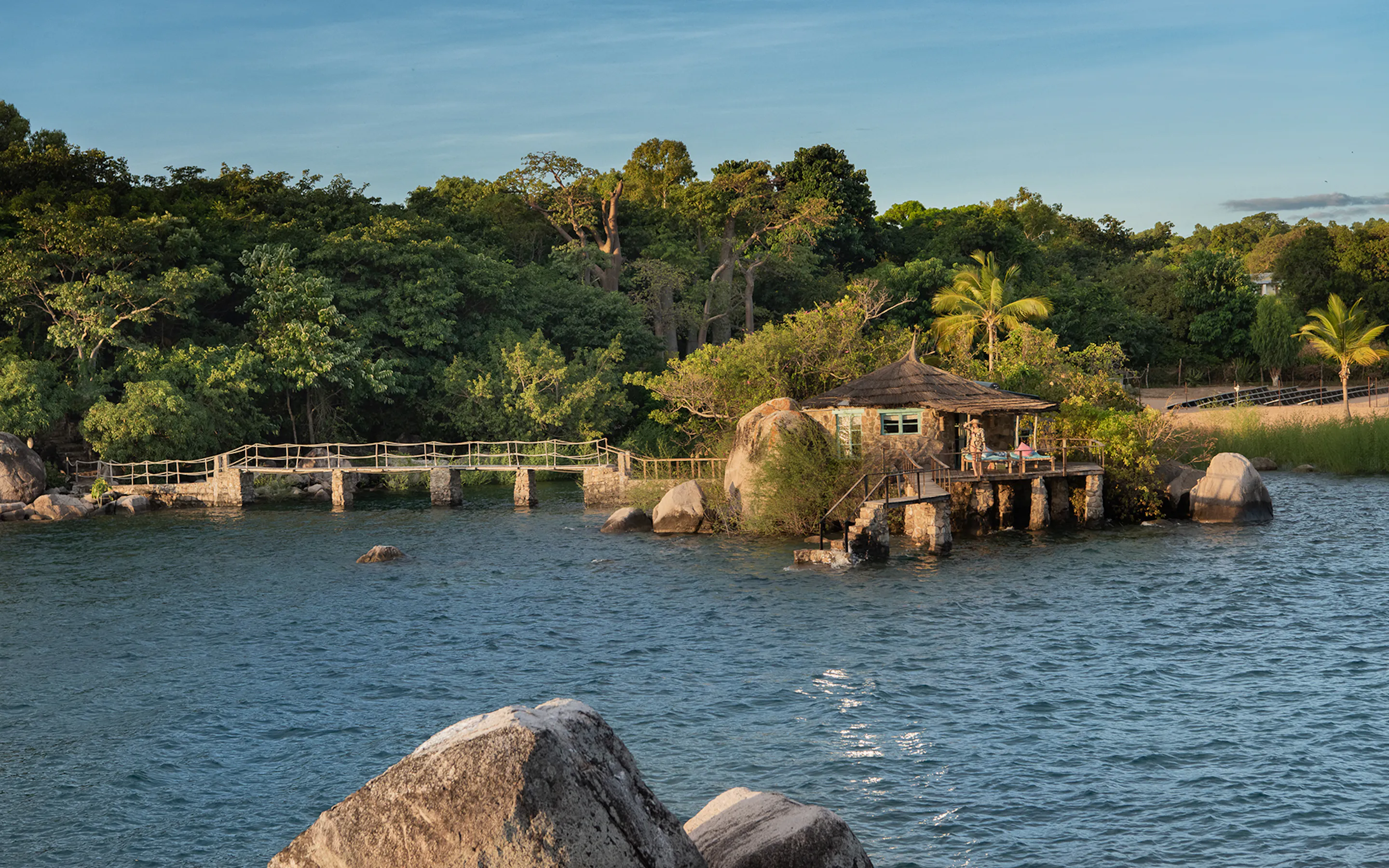 A lakeside pavilion sits above the water at Kaya Mawa, with boulders, palms, and lush island greenery behind.