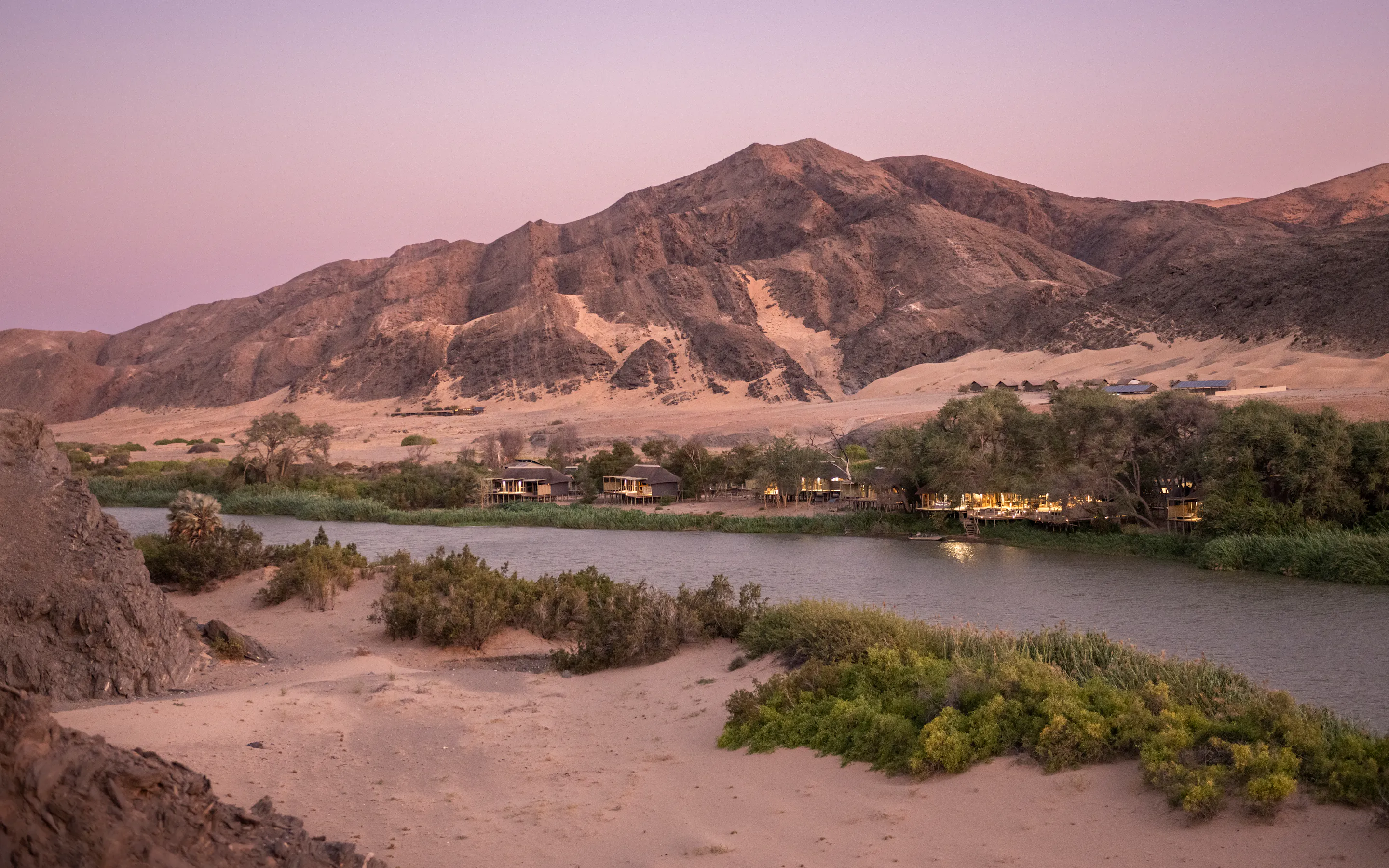 Serra Cafema sits beside the river at dusk, with rocky mountains rising behind green reeds and sandy banks.