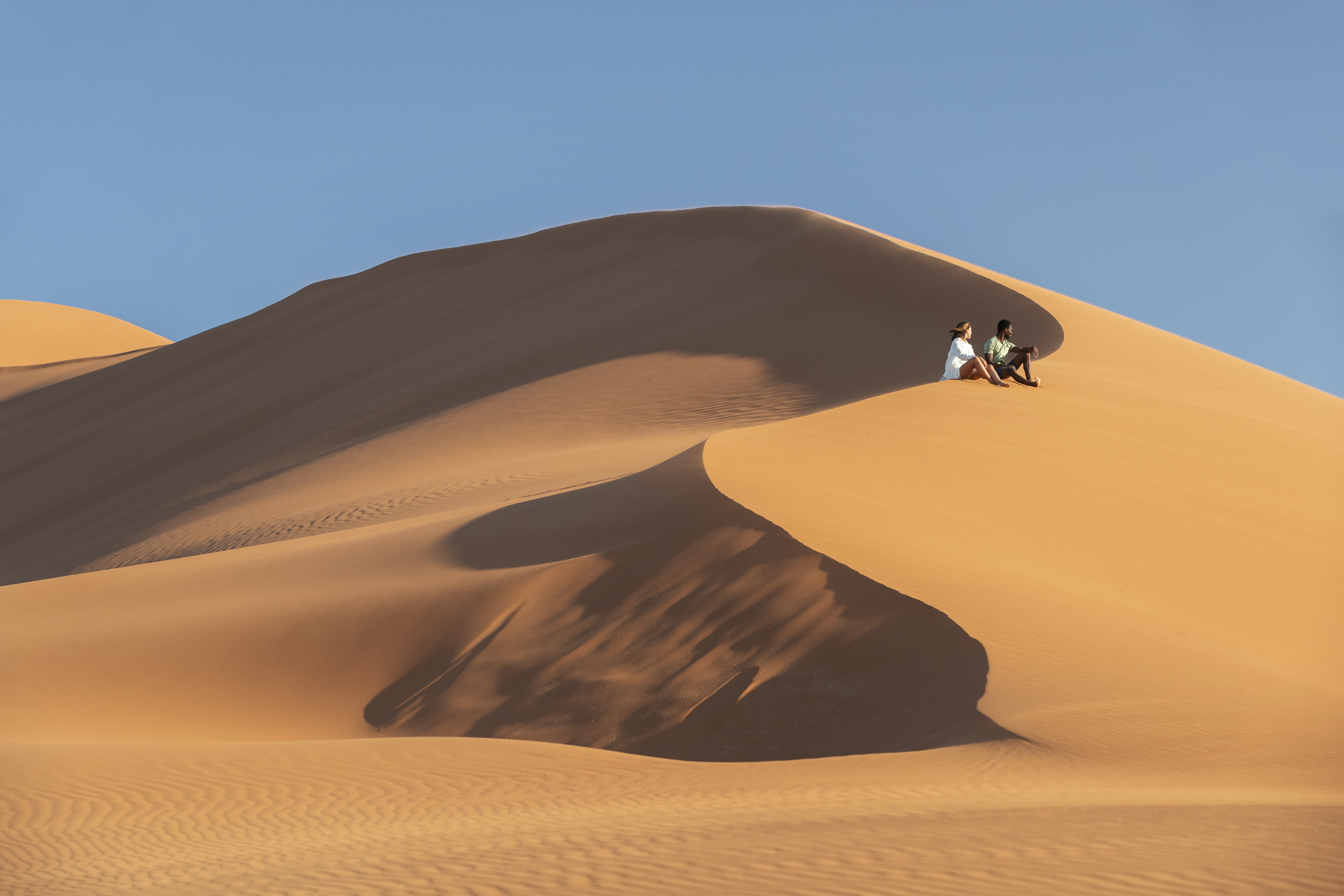 Two guests climb a sweeping sand dune near Kulala, with sharp shadows and blue sky above the ridge below.