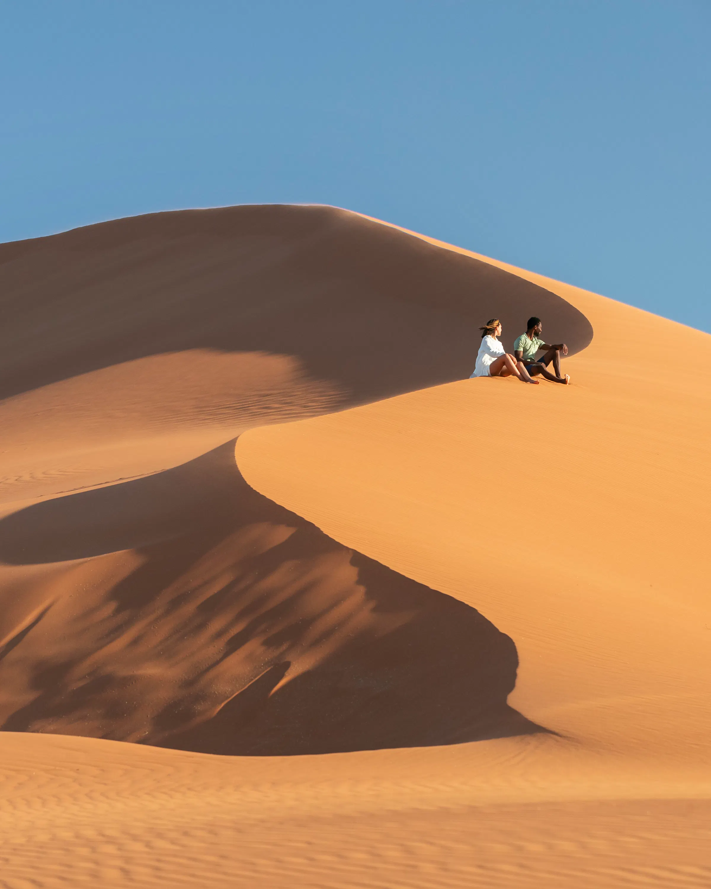 Two guests climb a sweeping sand dune near Kulala, with sharp shadows and blue sky above the ridge below.