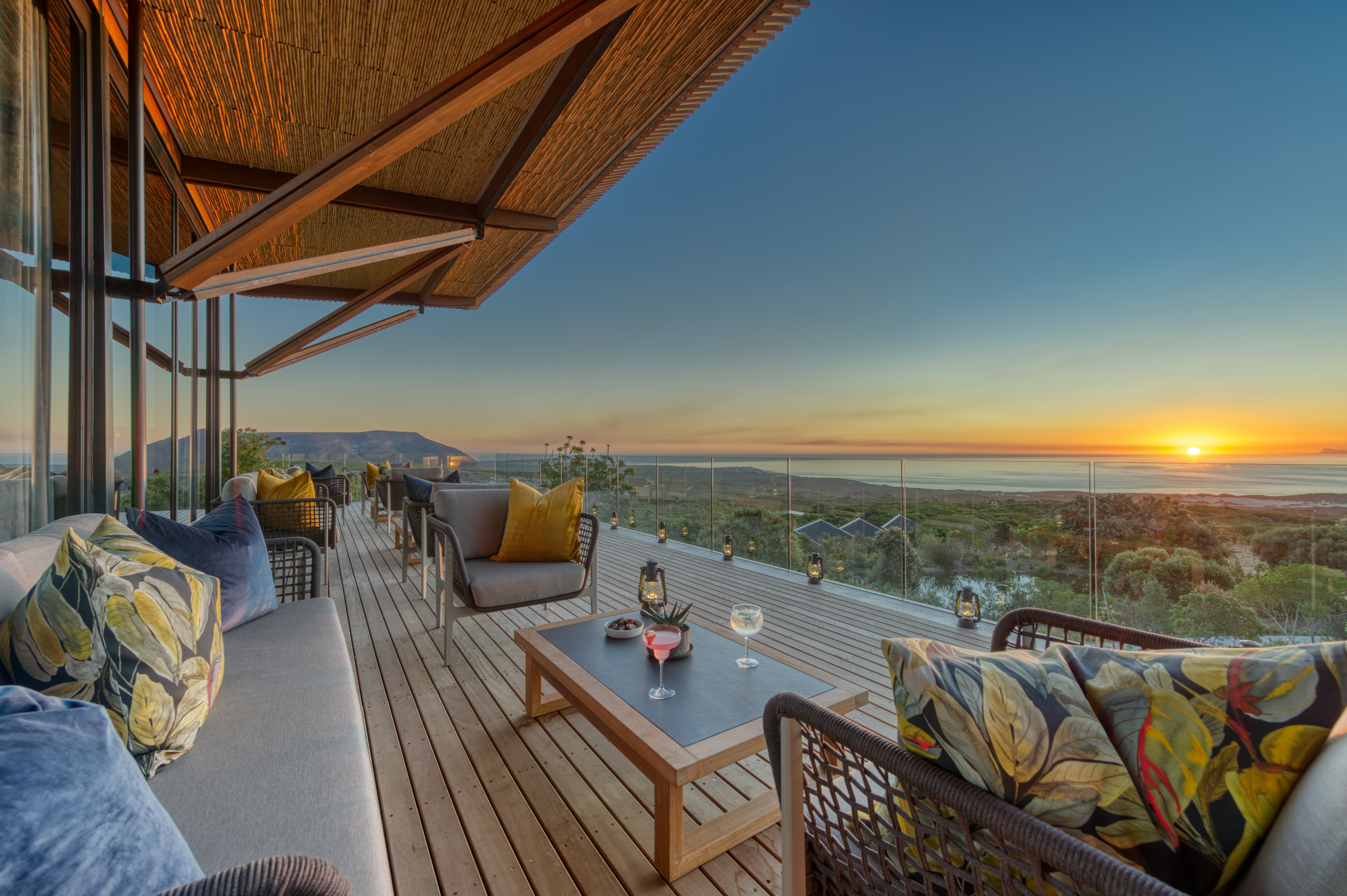 Cushioned chairs and sunset drinks line a glass-fronted deck at Grootbos, overlooking fynbos and the distant sea.