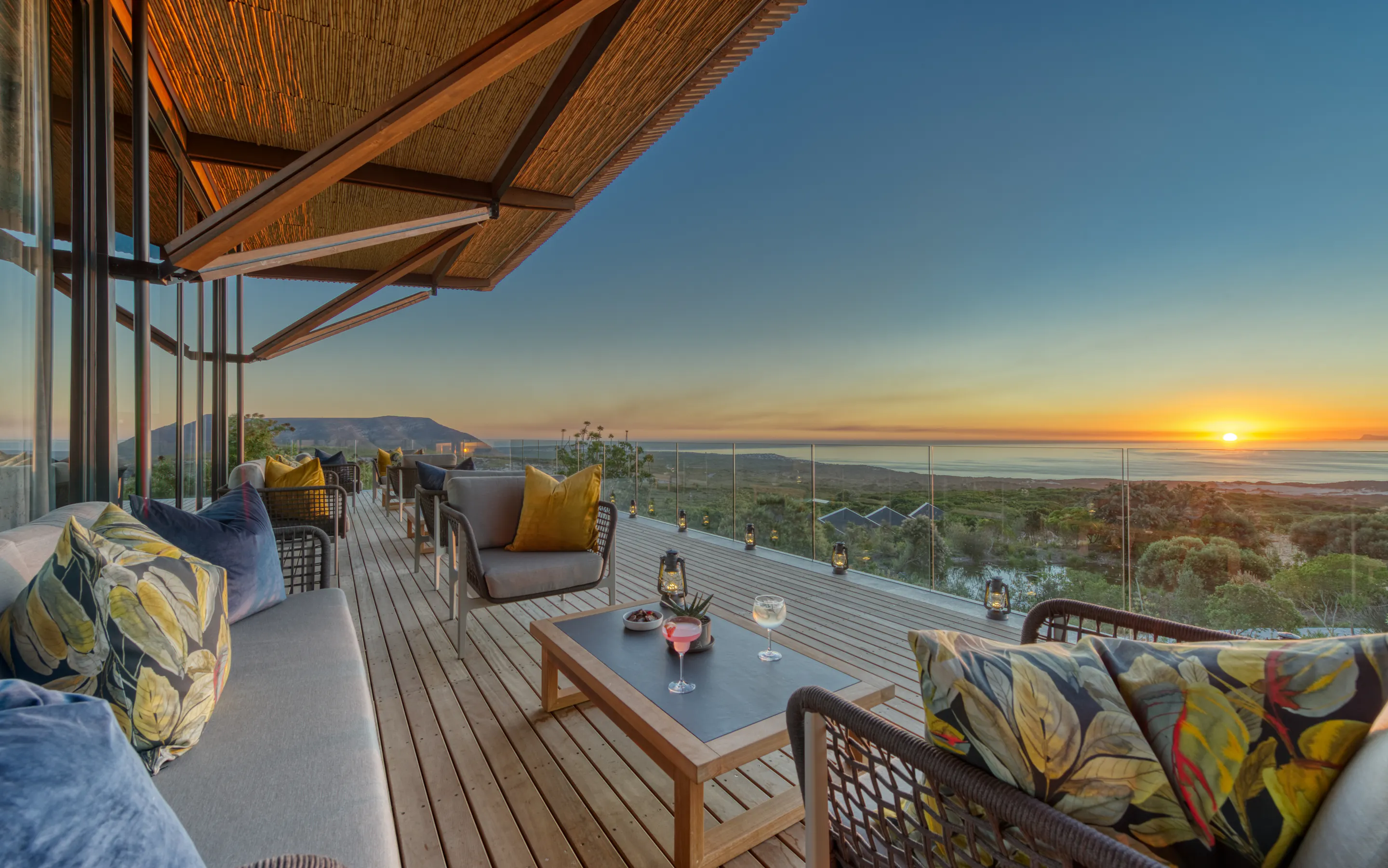Cushioned chairs and sunset drinks line a glass-fronted deck at Grootbos, overlooking fynbos and the distant sea.