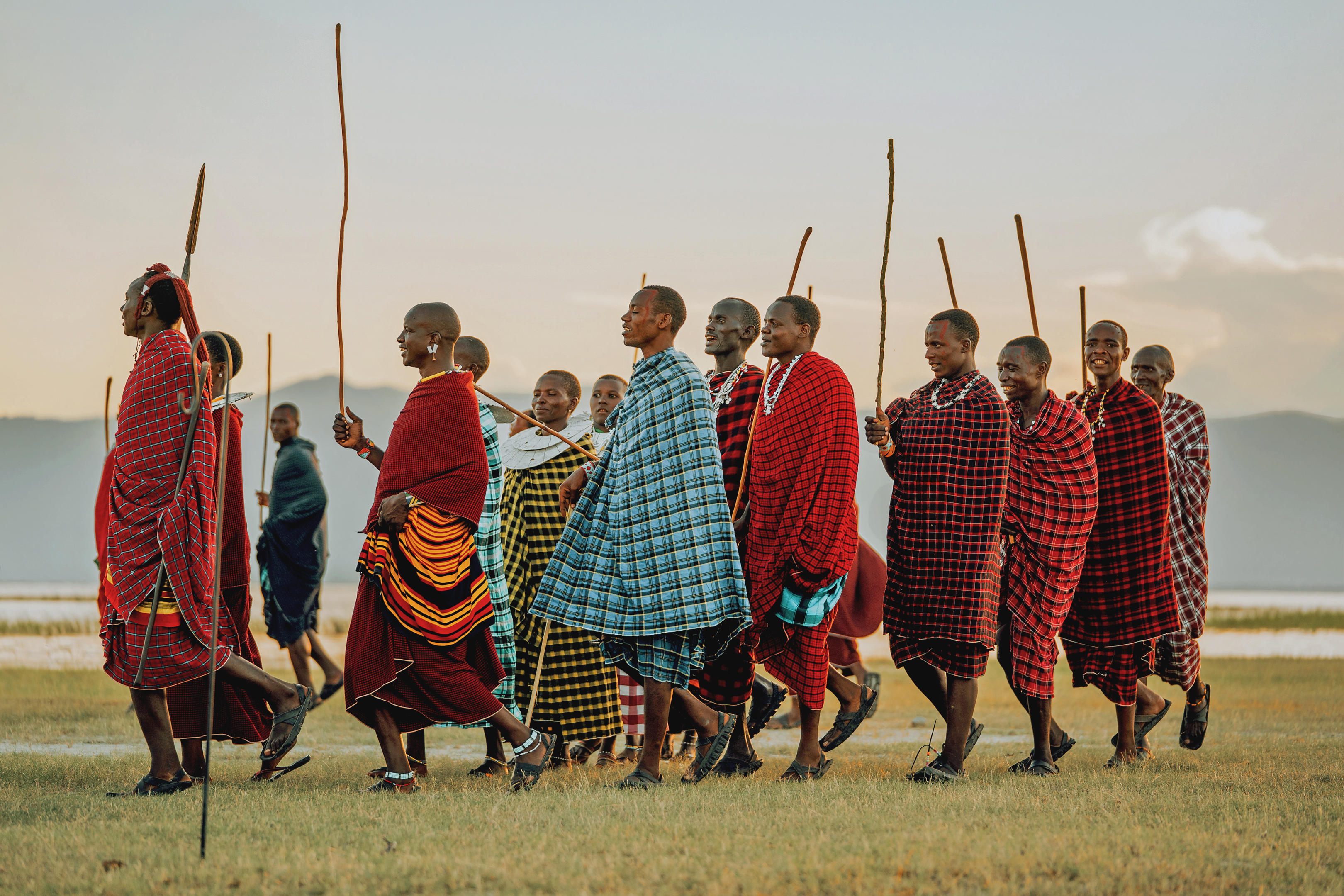 A line of Maasai men stands on open grass near Lake Manyara, wrapped in bright shukas against a pale sky.
