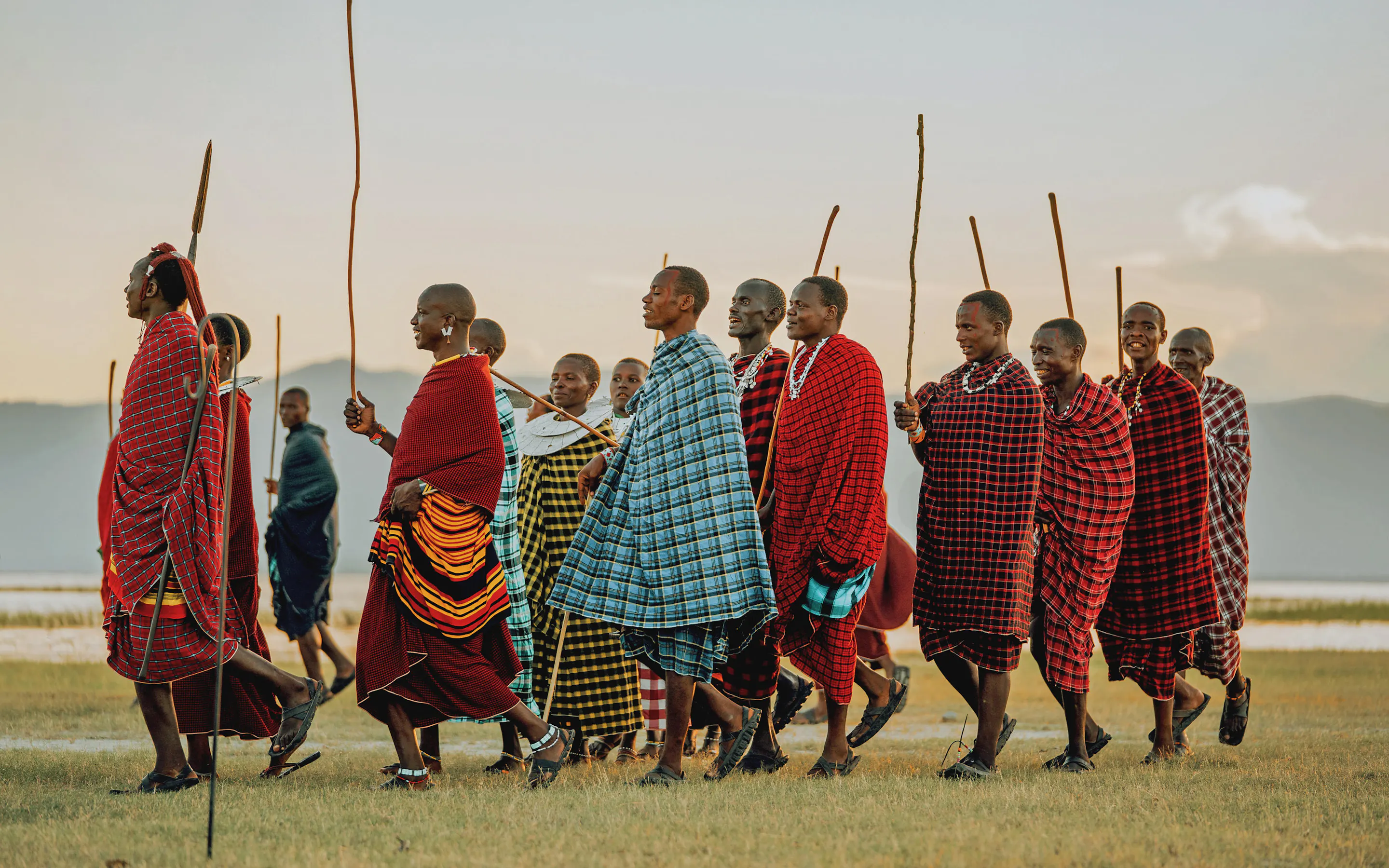 A line of Maasai men stands on open grass near Lake Manyara, wrapped in bright shukas against a pale sky.