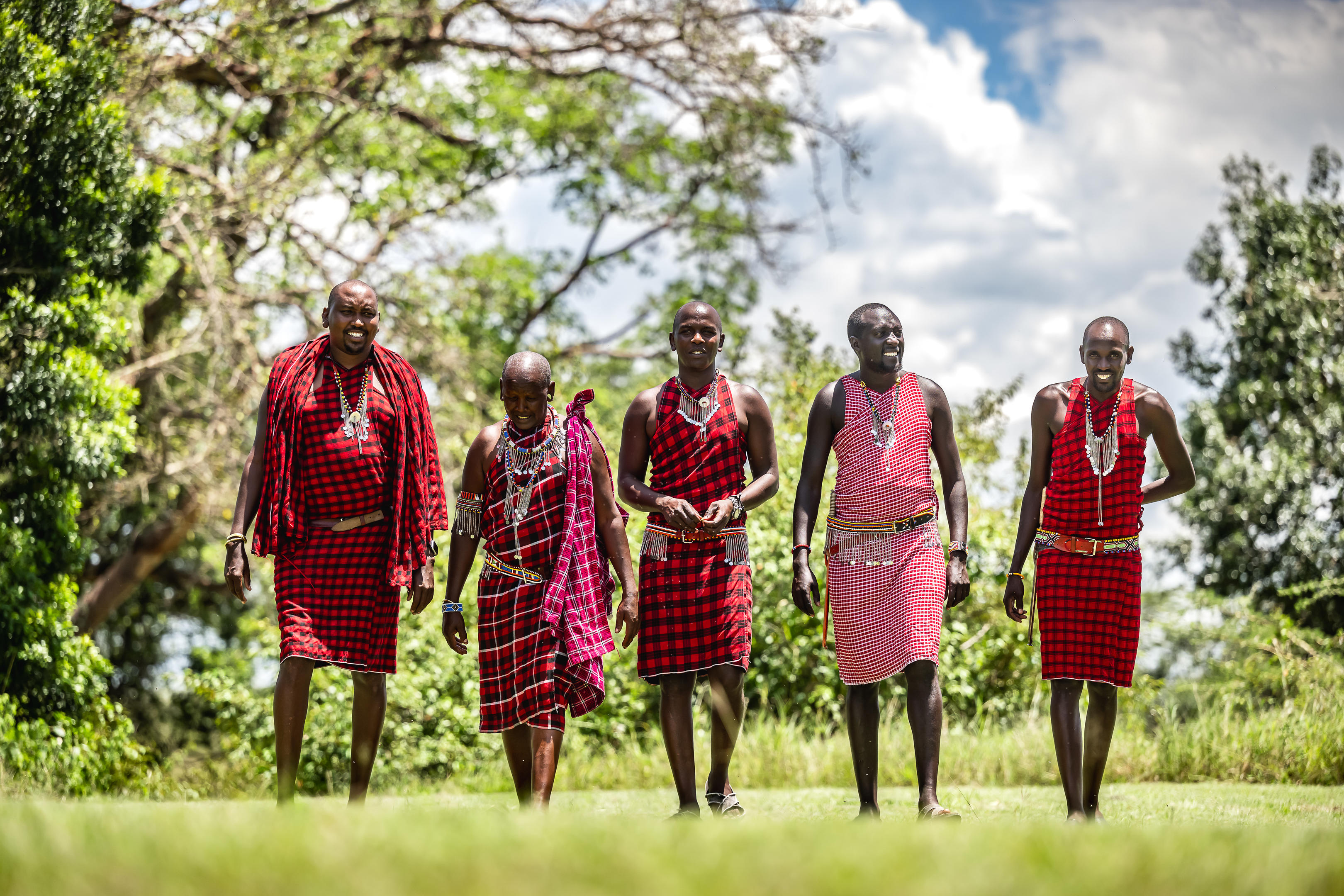 A group of Maasai stands together beneath trees, their red shukas bright against the green background behind.