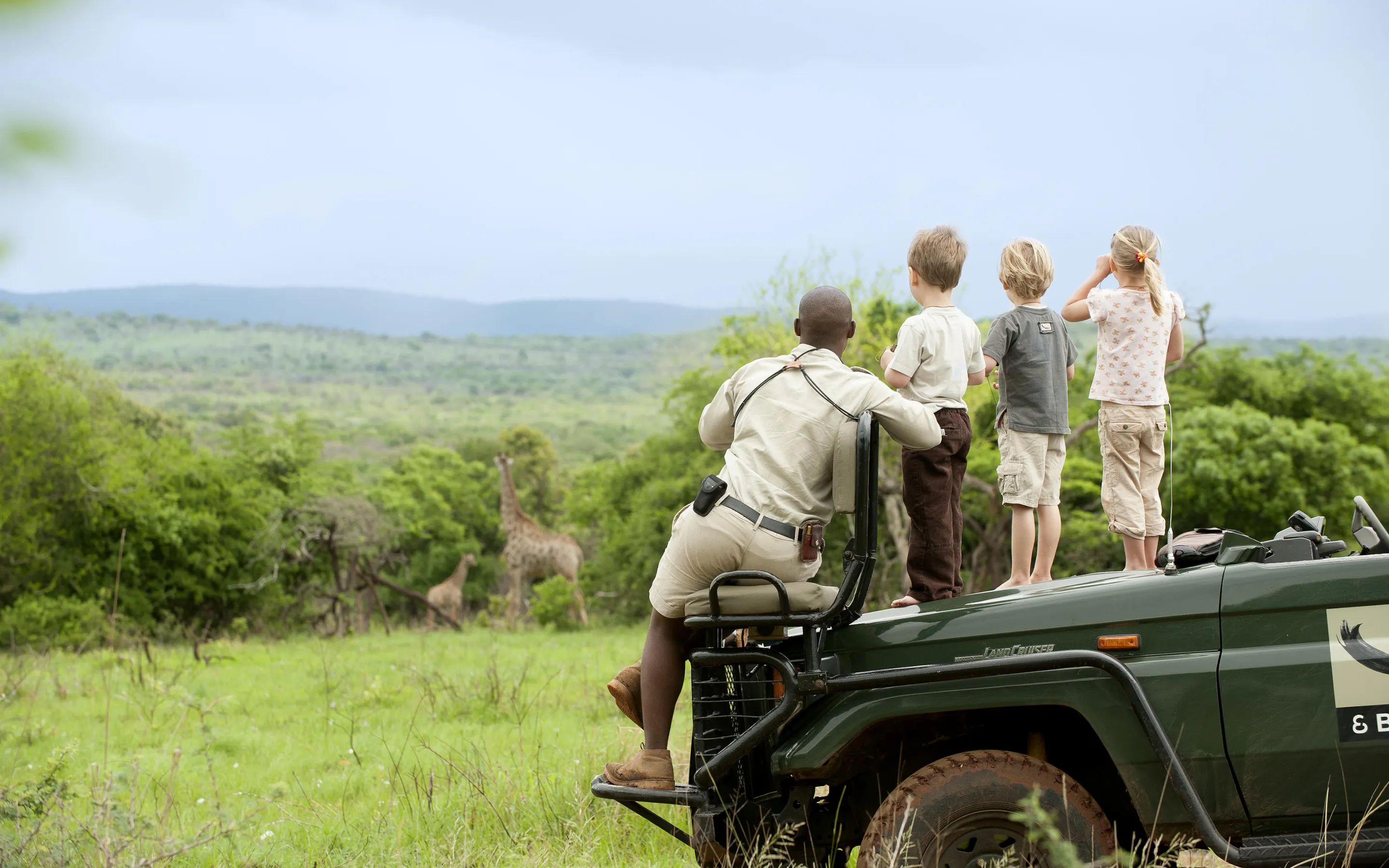 A ranger and three children stand on a safari vehicle watching giraffes, with green hills stretching beyond them.