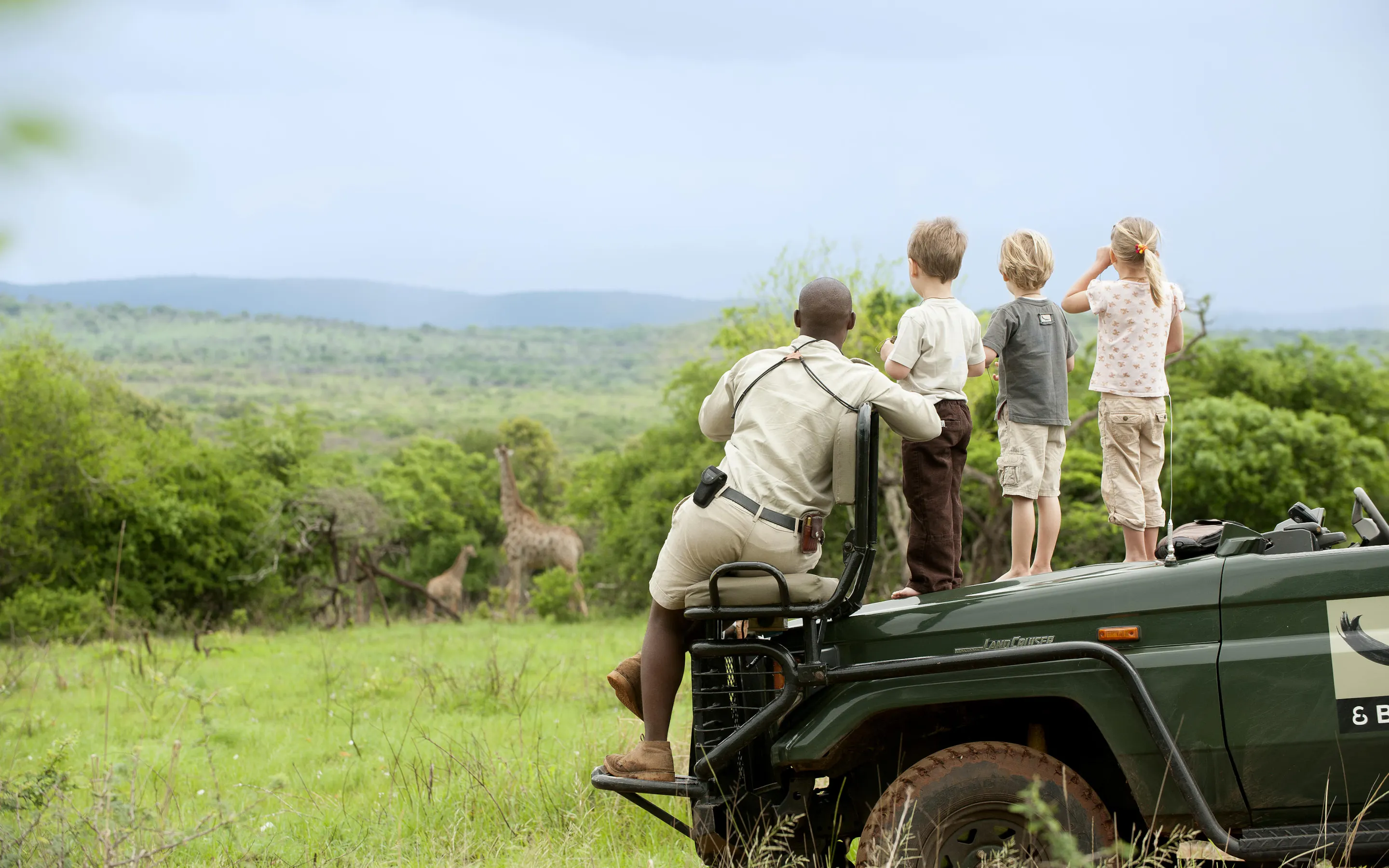 A ranger and three children stand on a safari vehicle watching giraffes, with green hills stretching beyond them.