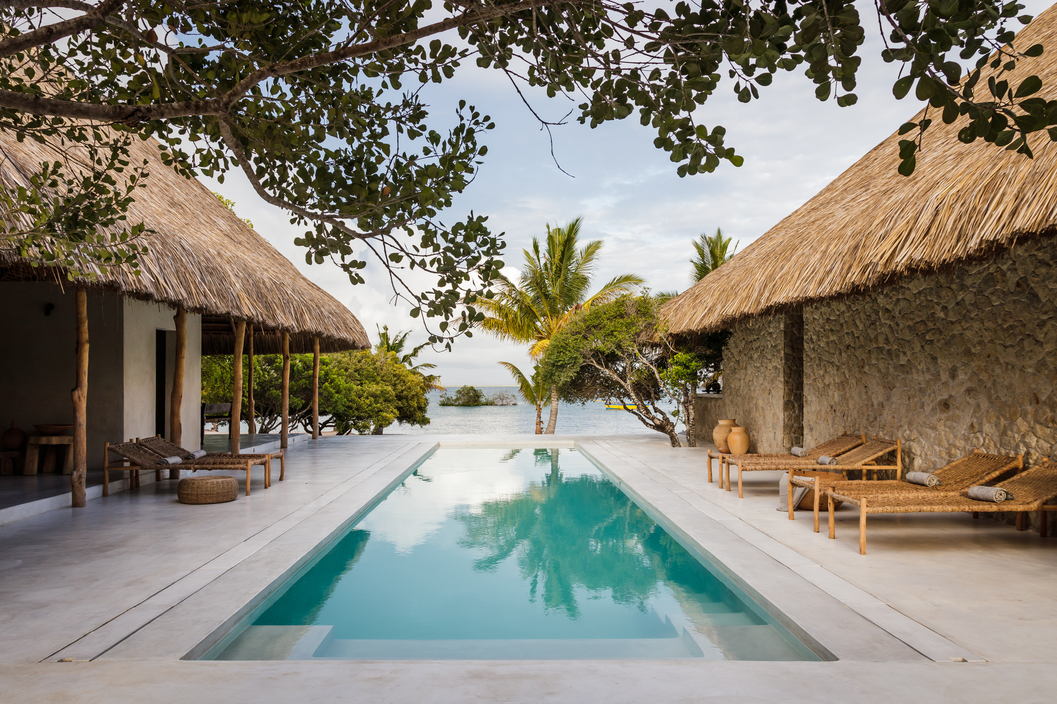 A long pool leads toward the sea at Sussurro, framed by thatched villas, loungers, and coastal trees beyond.
