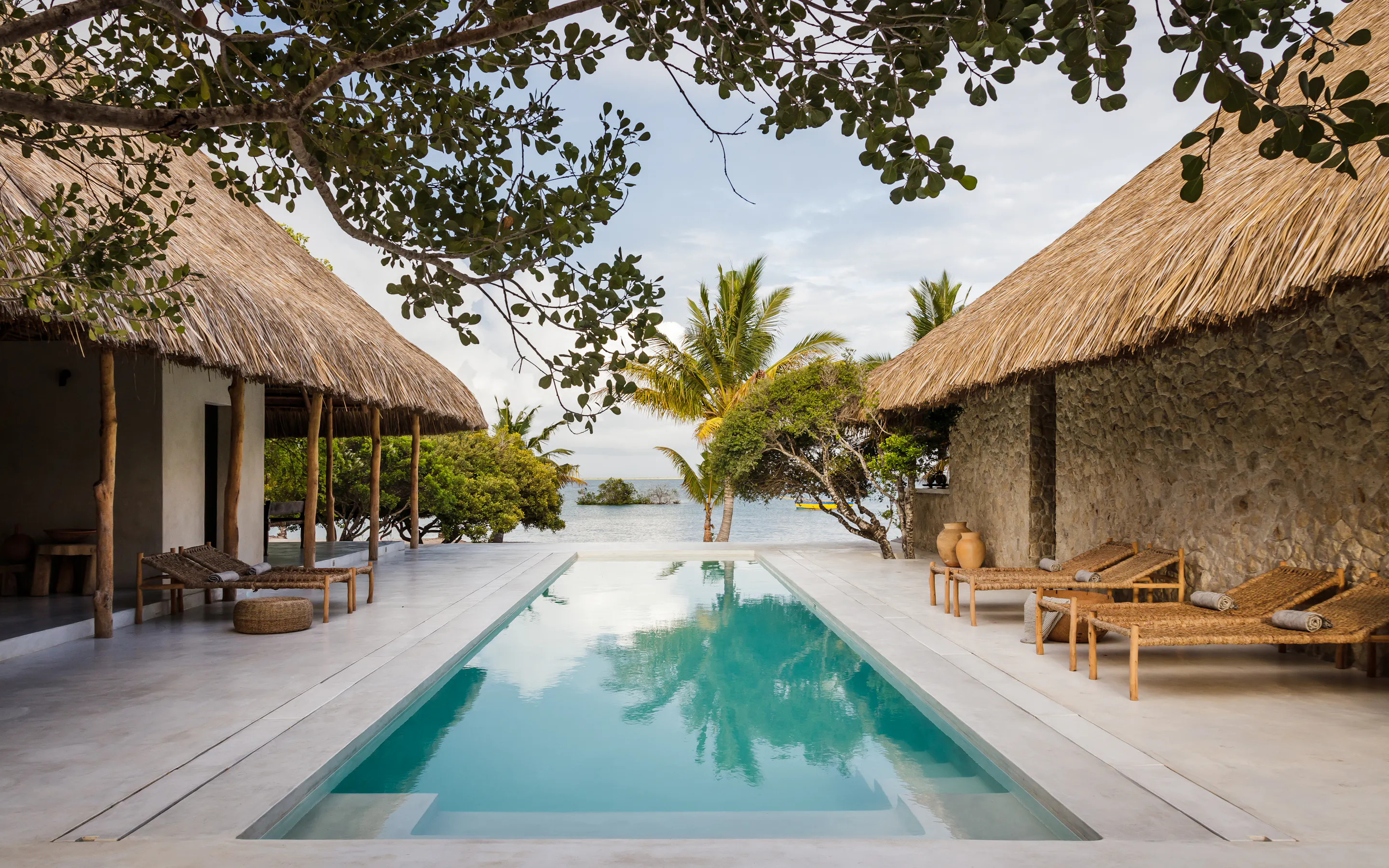 A long pool leads toward the sea at Sussurro, framed by thatched villas, loungers, and coastal trees beyond.