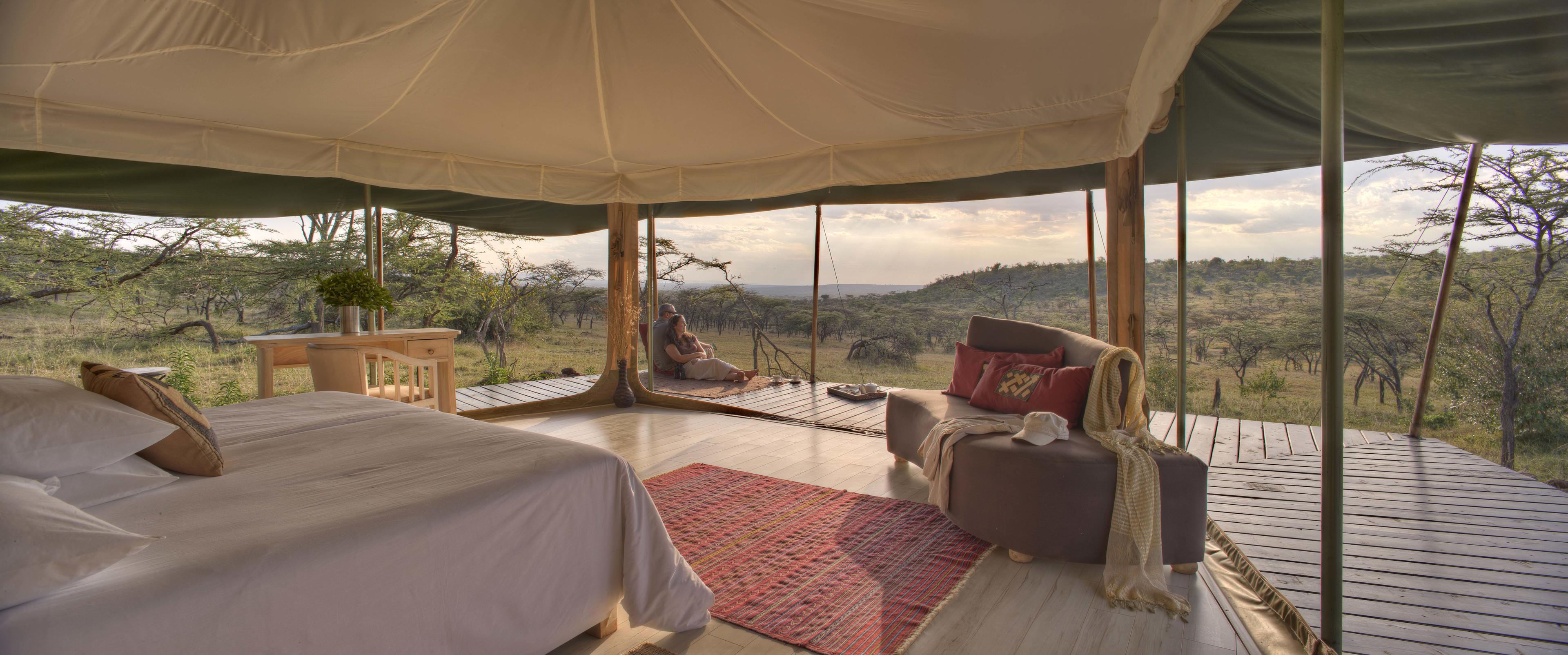 A tented room at Kicheche Valley Camp looks out over the hills, with a bed, rug, and deck in warm evening light.