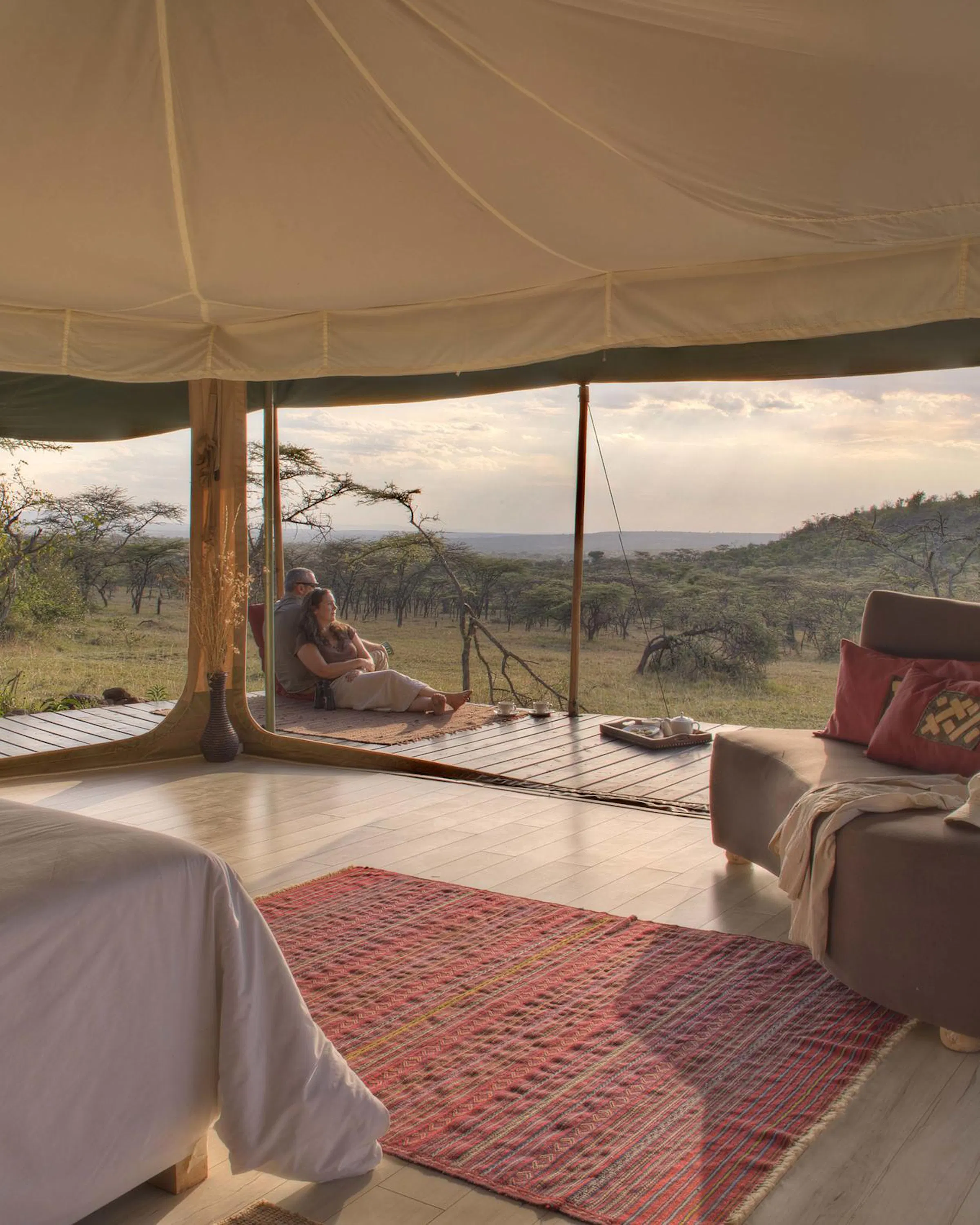 A tented room at Kicheche Valley Camp looks out over the hills, with a bed, rug, and deck in warm evening light.