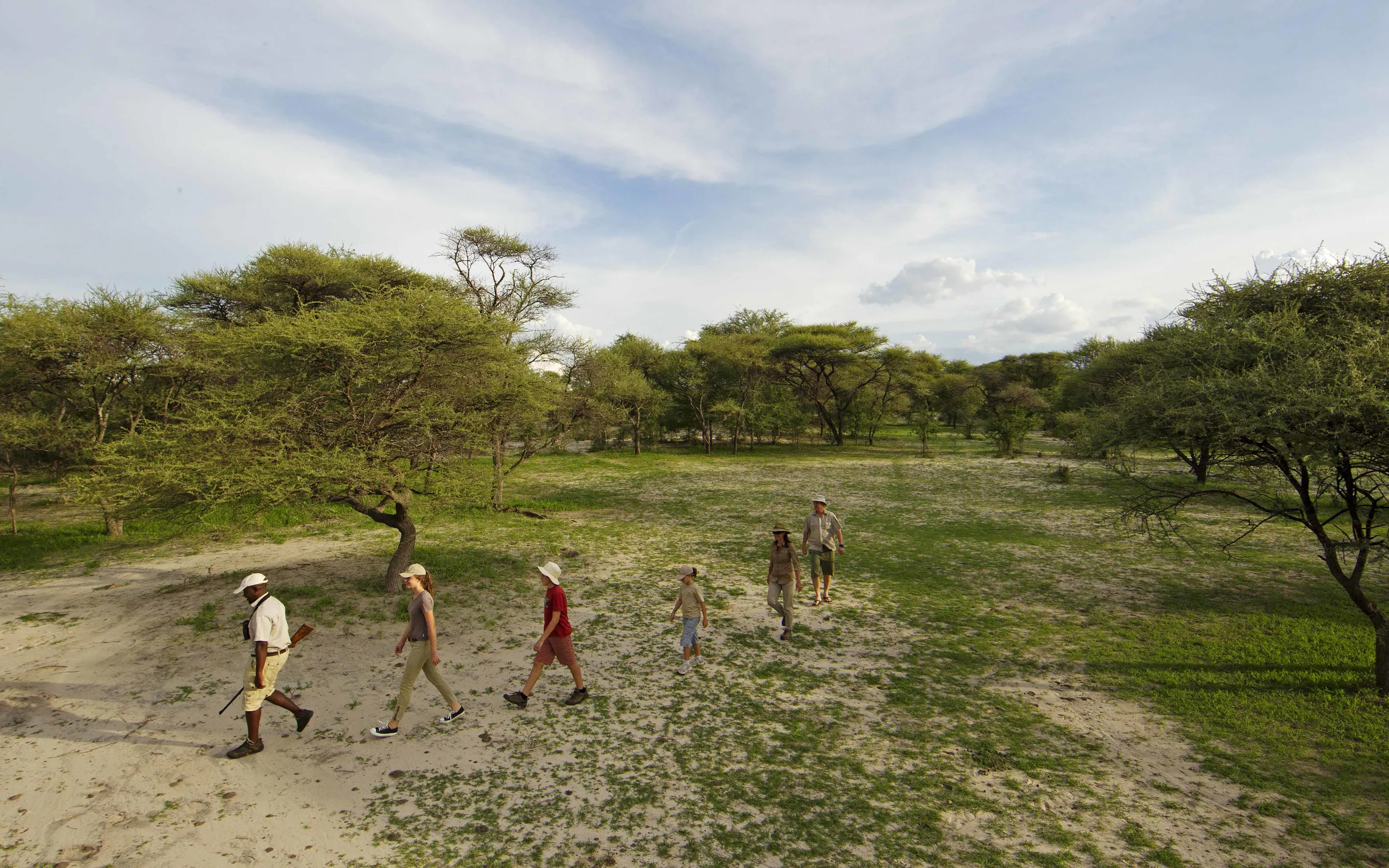 A guide leads a family walking safari across a sandy clearing, with acacia trees and wide sky across the delta.