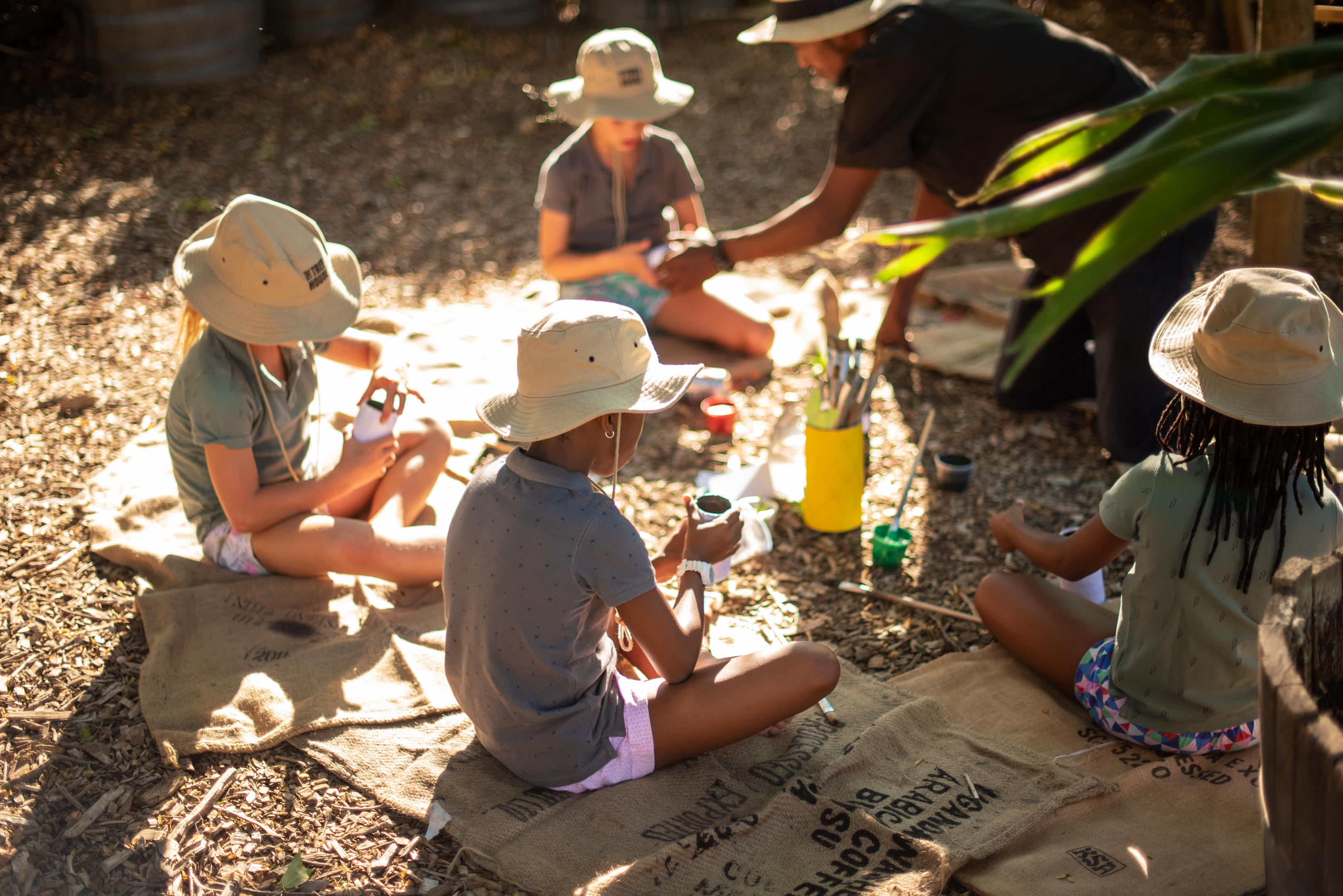 Children sit in a circle during an outdoor activity at Boschendal, with hats, jars, and craft materials on the mat.