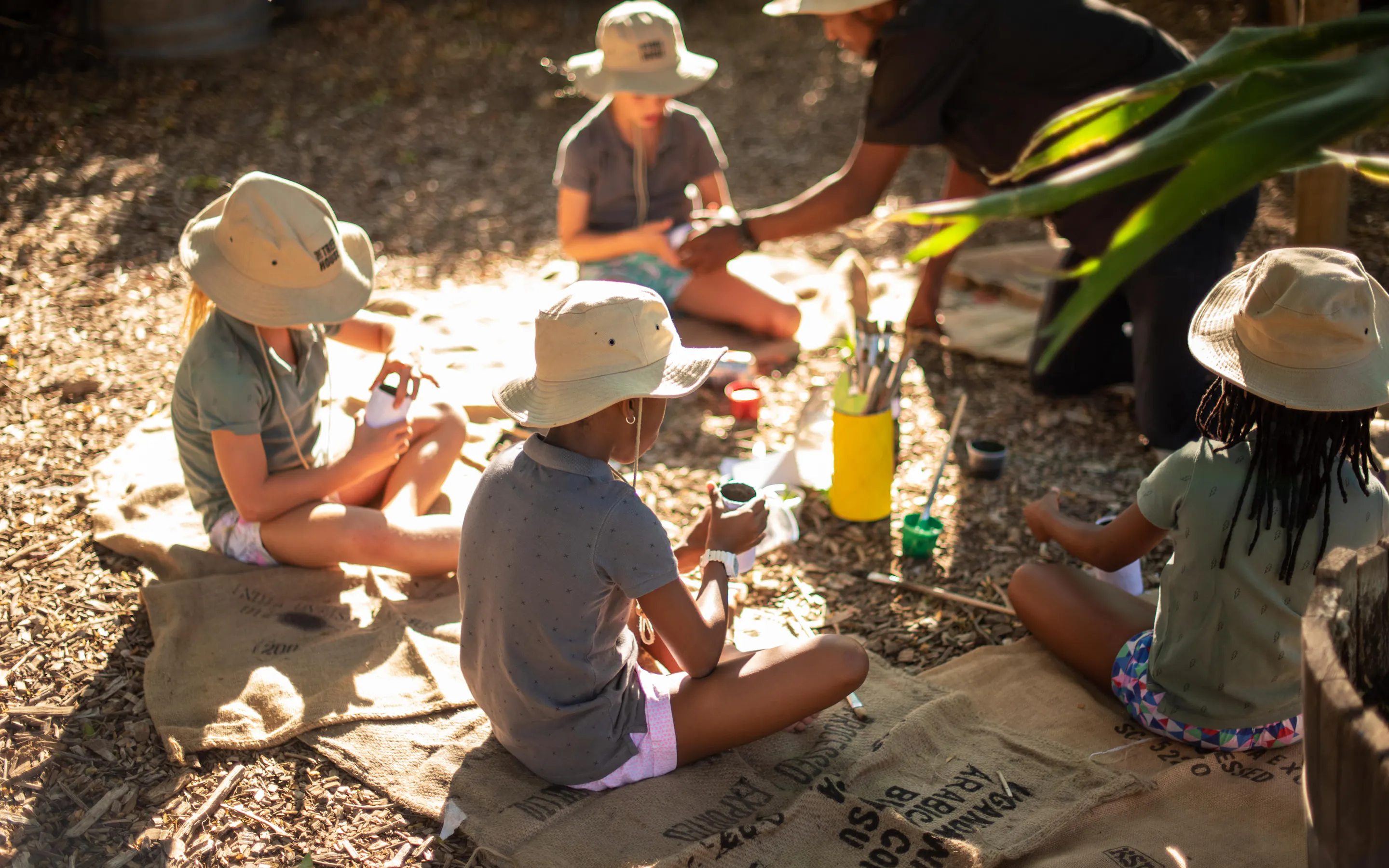 Children sit in a circle during an outdoor activity at Boschendal, with hats, jars, and craft materials on the mat.