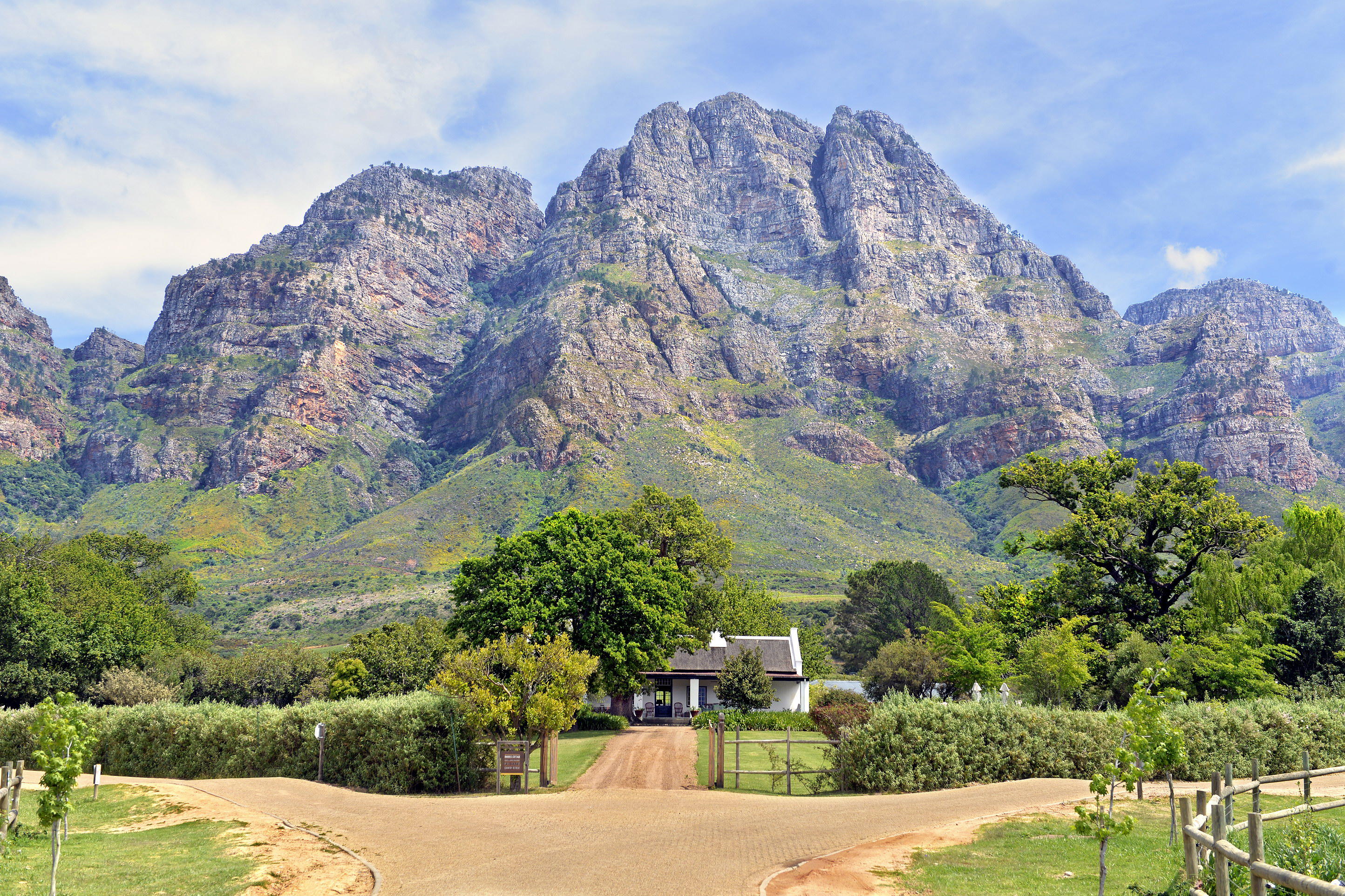 A farm road leads toward Boschendal beneath dramatic Franschhoek mountains, with trees and vines in the foreground.
