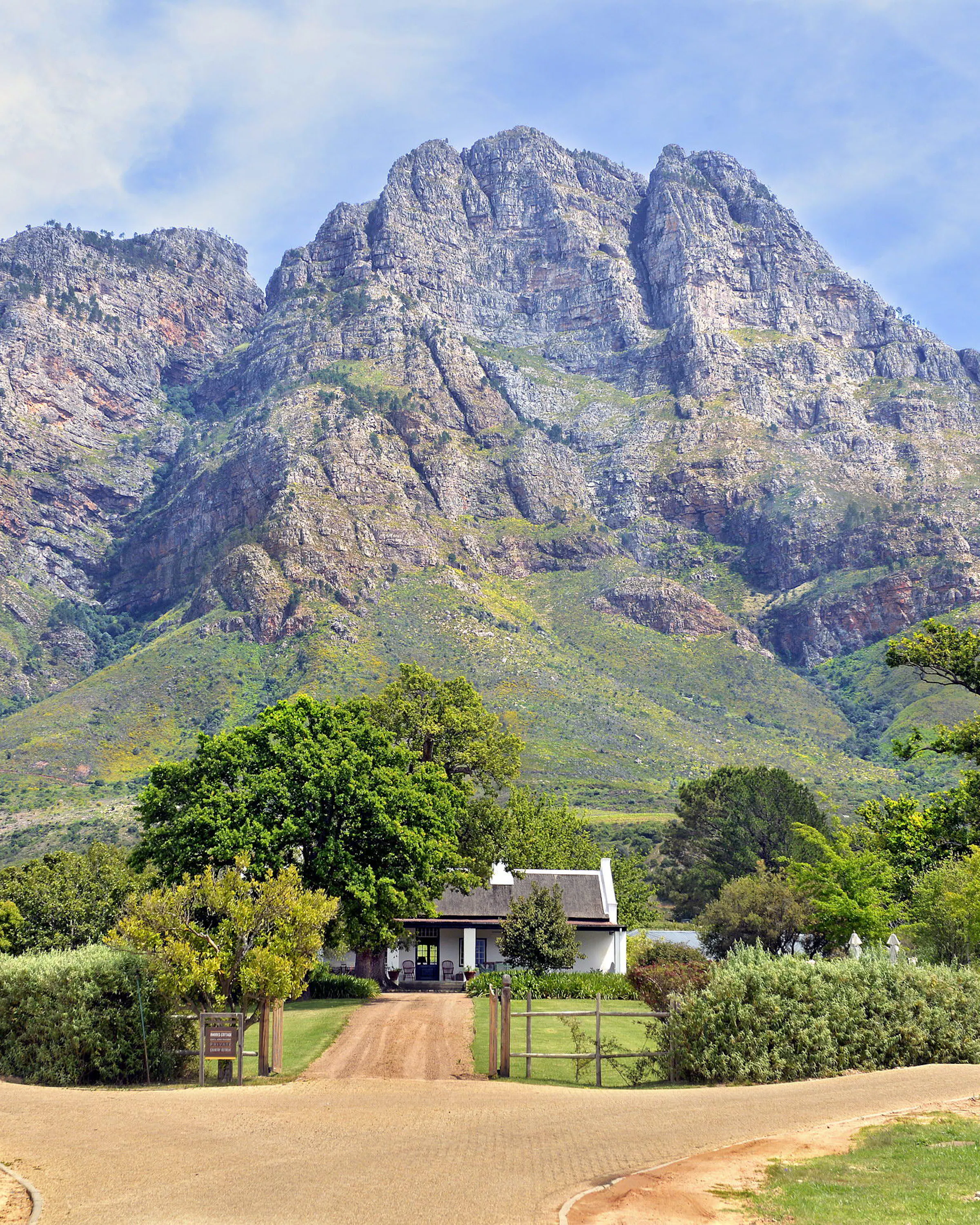 A farm road leads toward Boschendal beneath dramatic Franschhoek mountains, with trees and vines in the foreground.