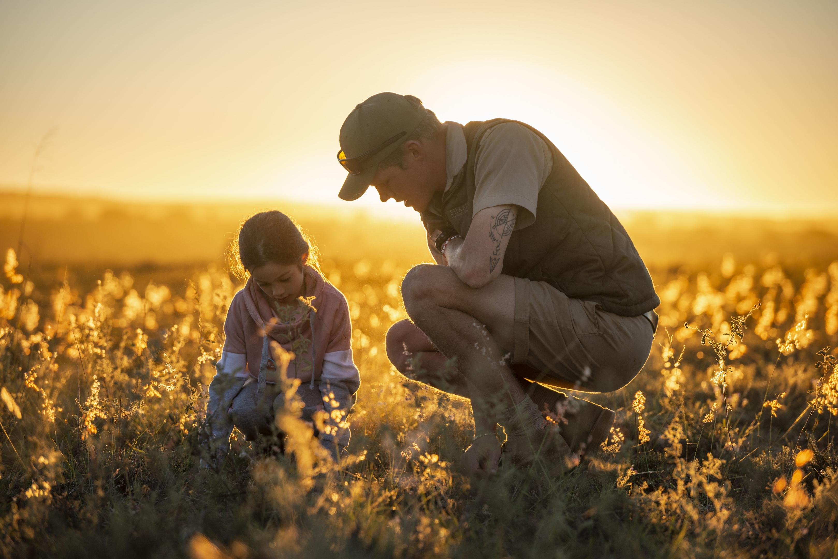 An adult and child crouch in golden grass at sunset in Shamwari, studying something small on the ground together.