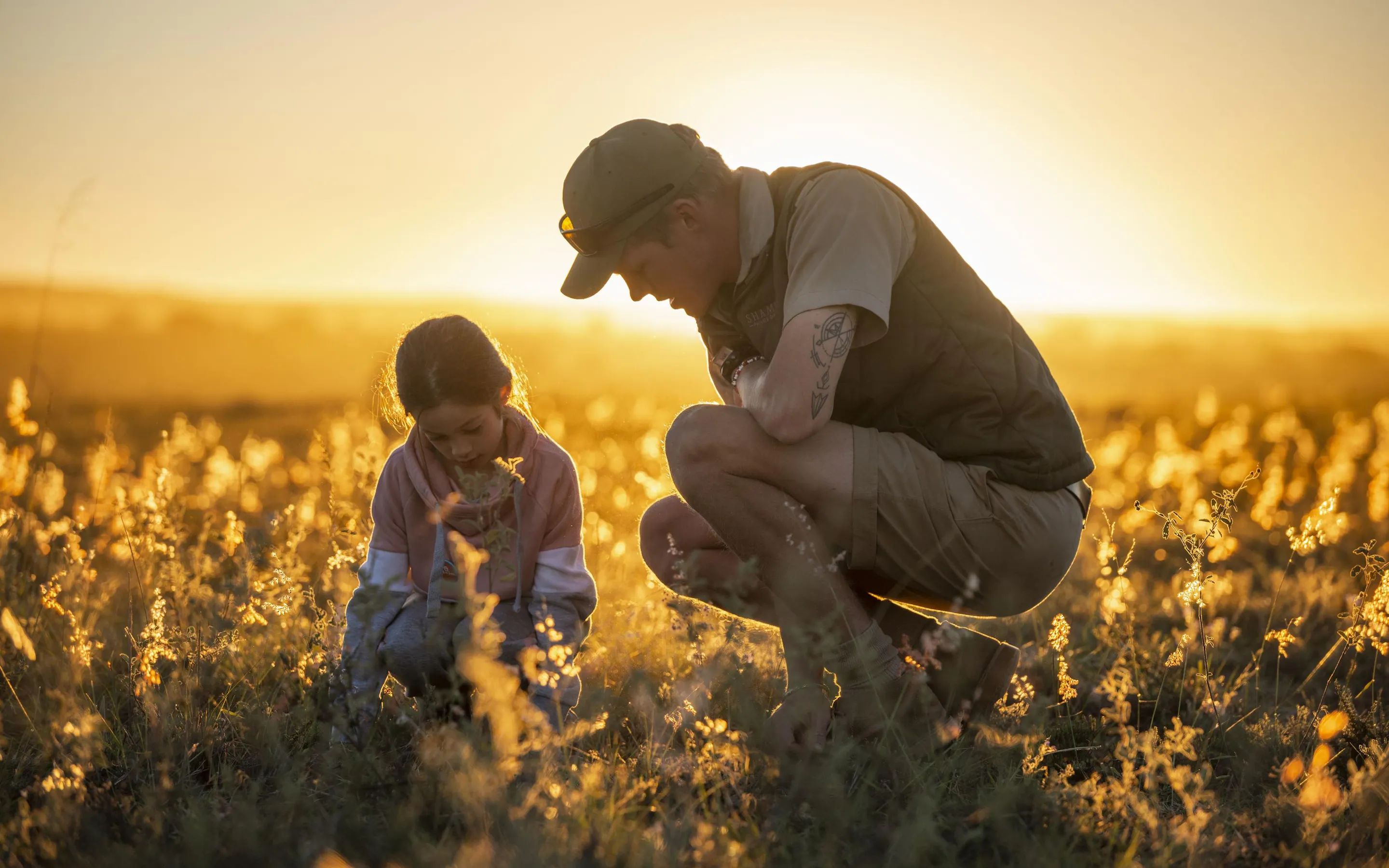 An adult and child crouch in golden grass at sunset in Shamwari, studying something small on the ground together.