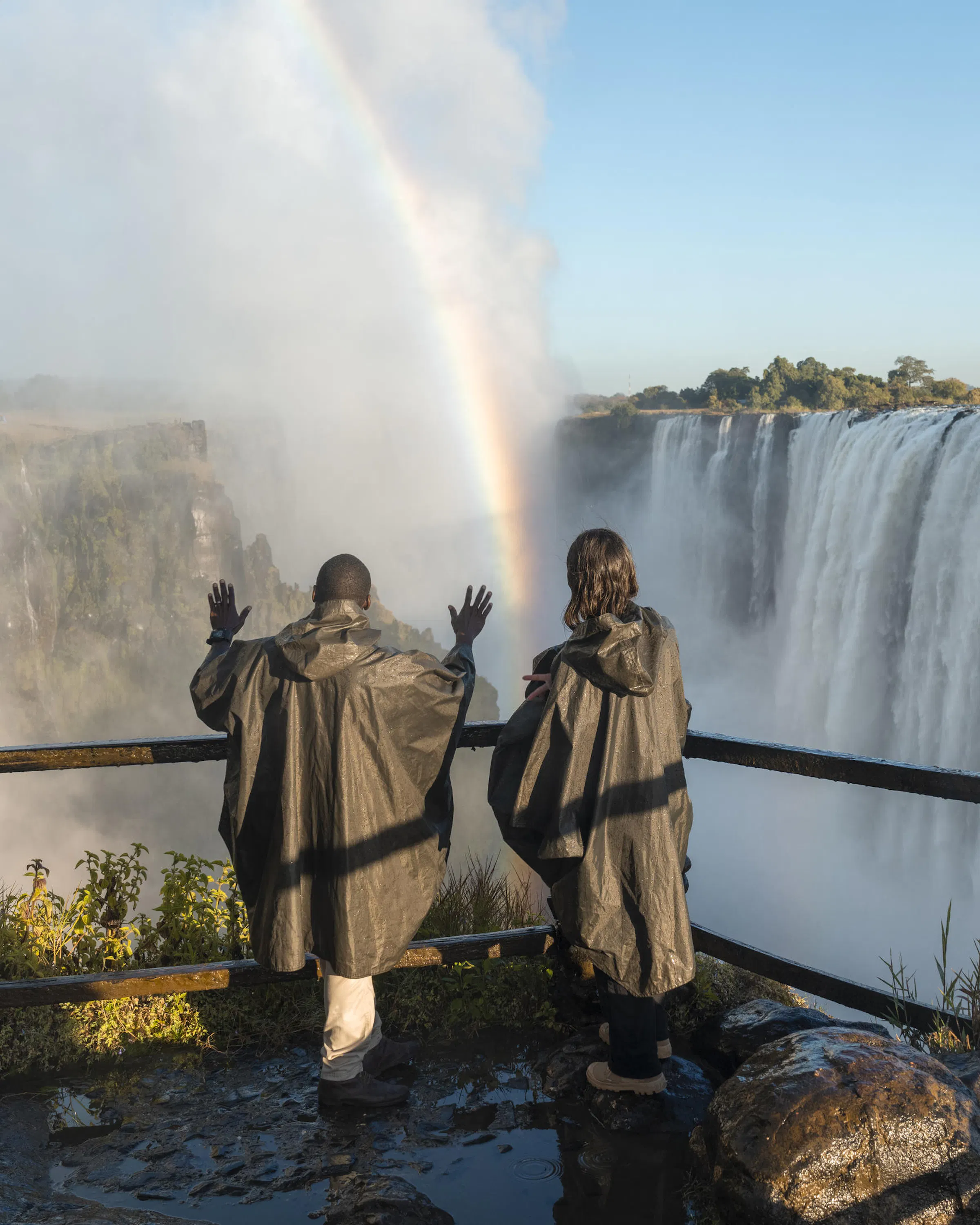 Visitors in ponchos stand above Victoria Falls on Livingstone Island, with spray rising and a rainbow over the gorge.