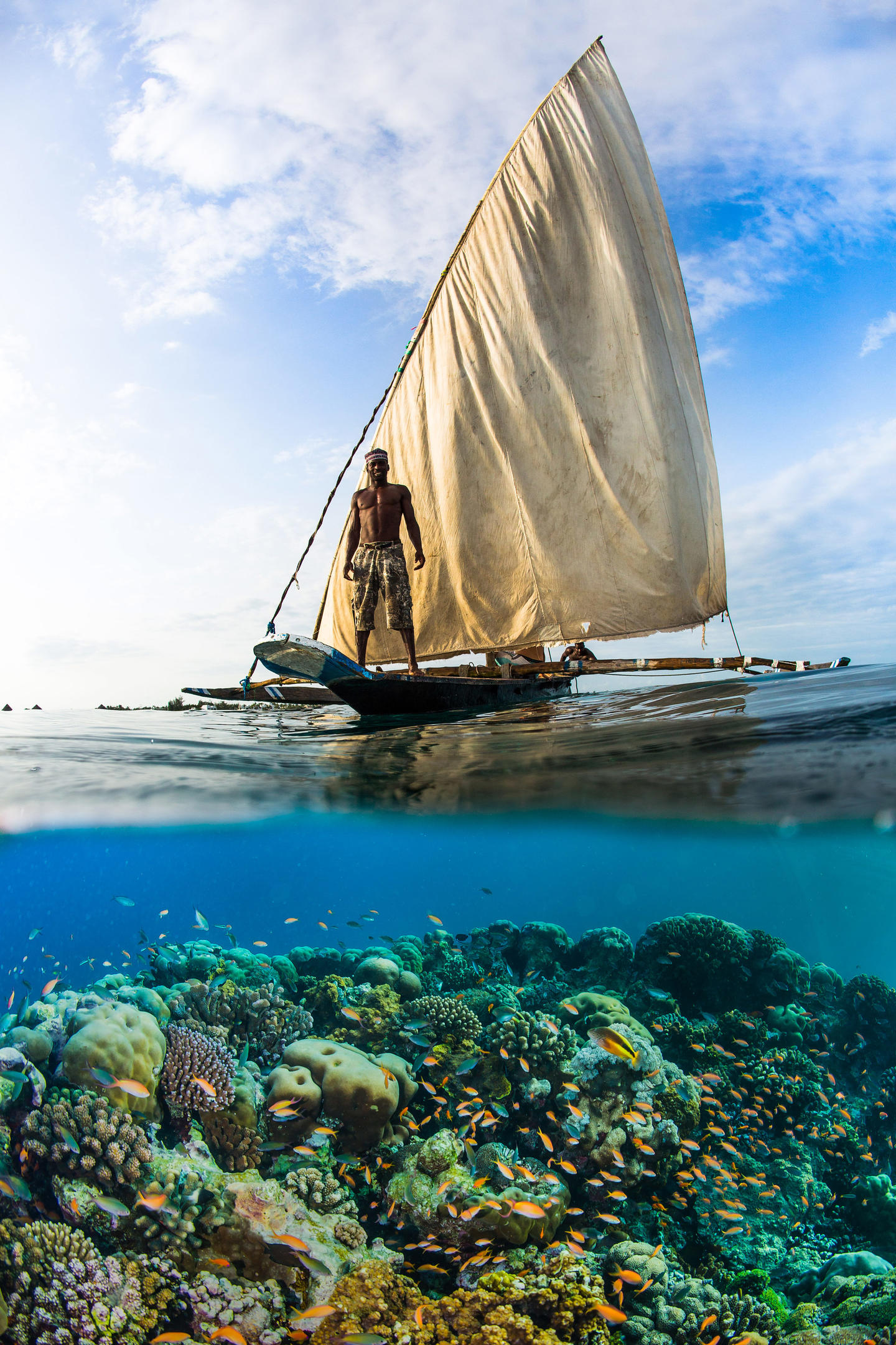 A traditional ngalawa sailboat glides above a coral reef off Pemba, with the sailor framed against bright sky.