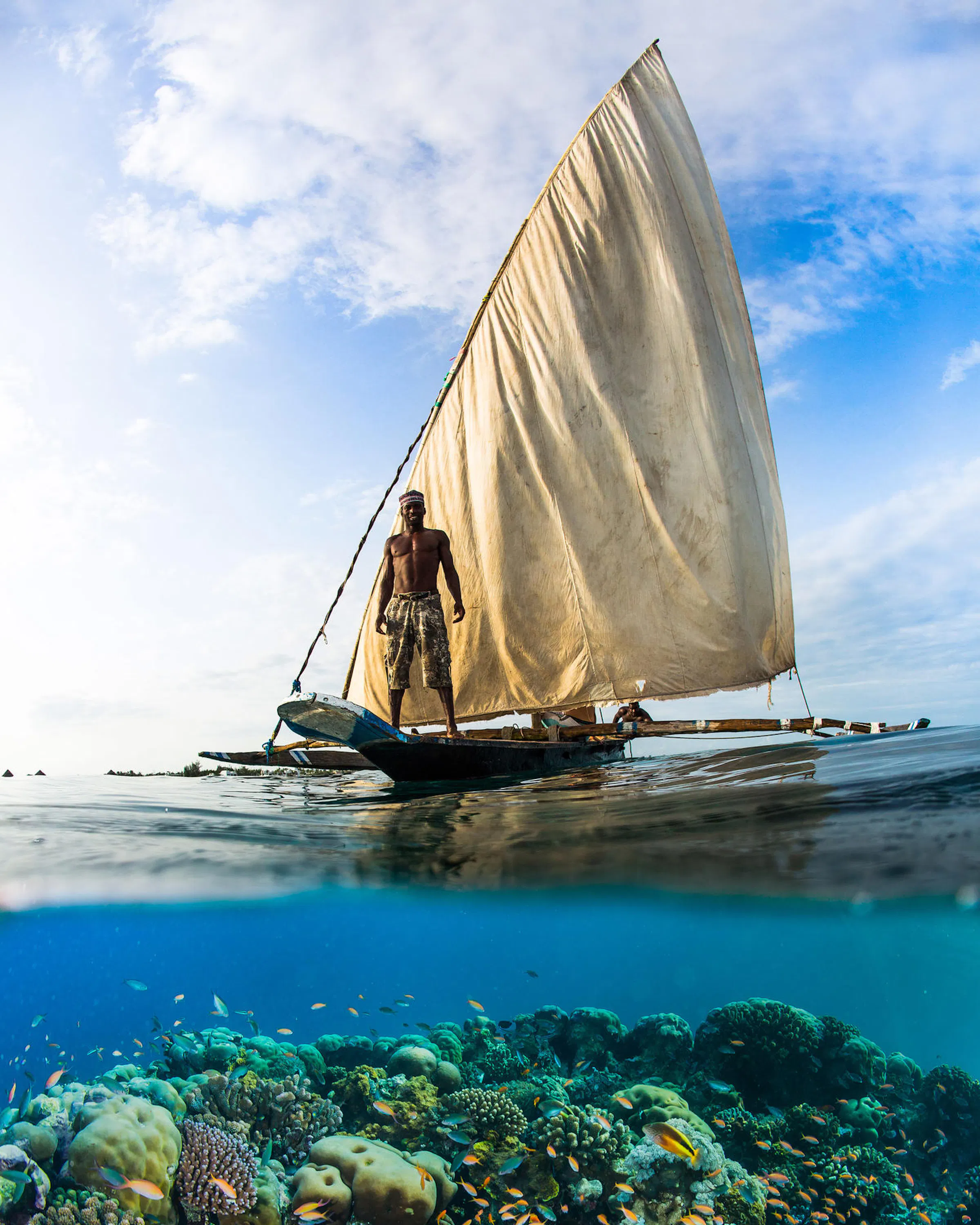 A traditional ngalawa sailboat glides above a coral reef off Pemba, with the sailor framed against bright sky.