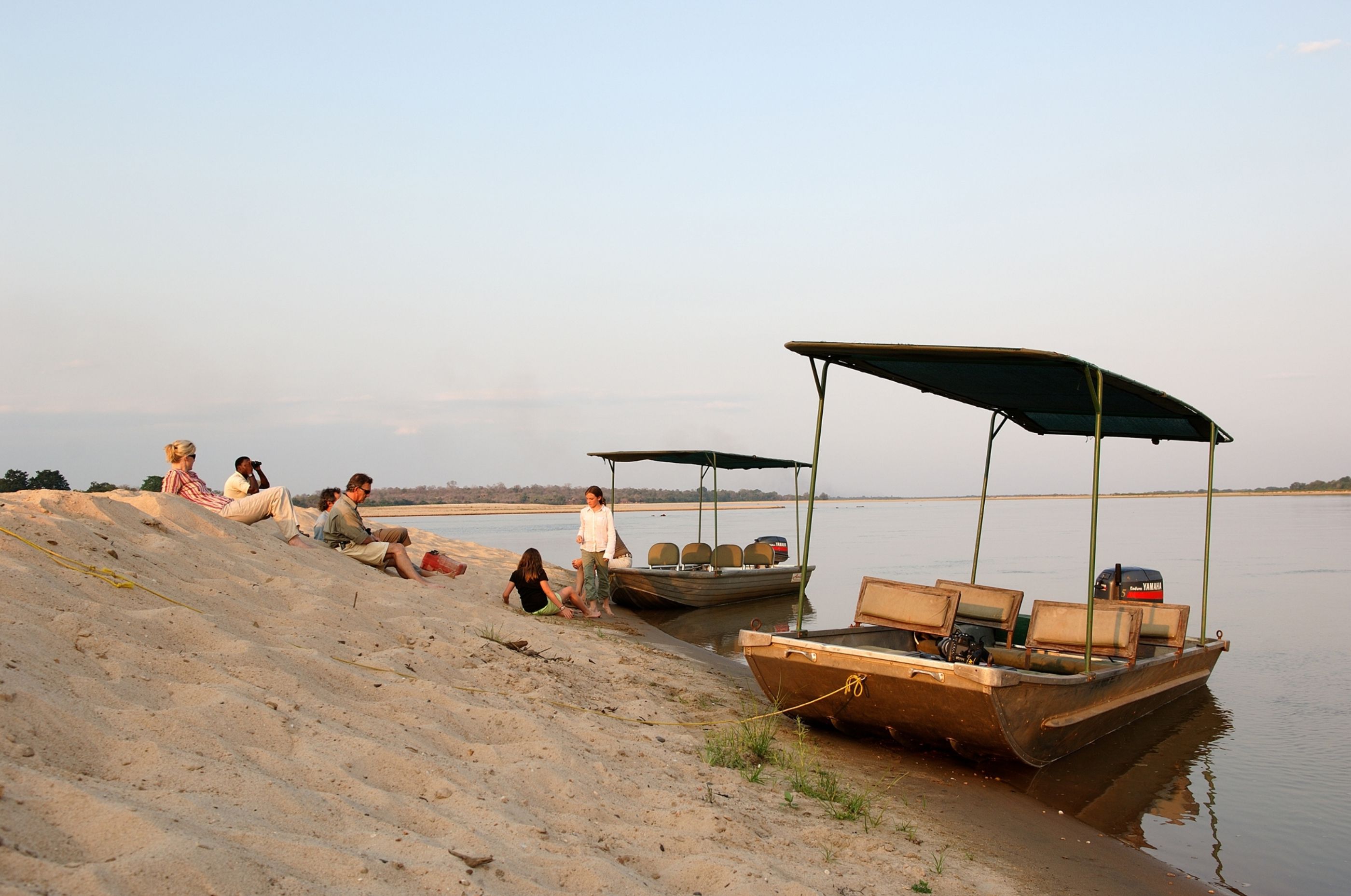 Two safari boats rest on a sandy riverbank in Nyerere as guests sit nearby, with still water and reeds beside them.