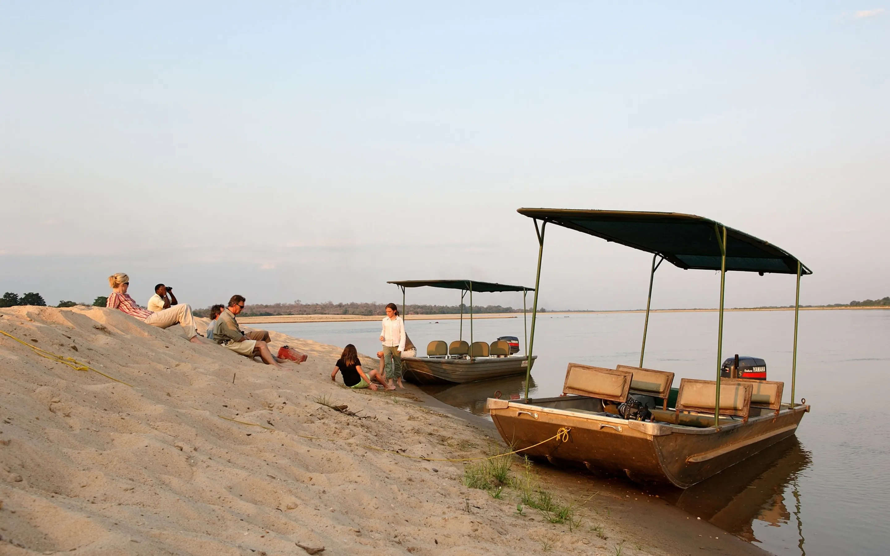 Two safari boats rest on a sandy riverbank in Nyerere as guests sit nearby, with still water and reeds beside them.